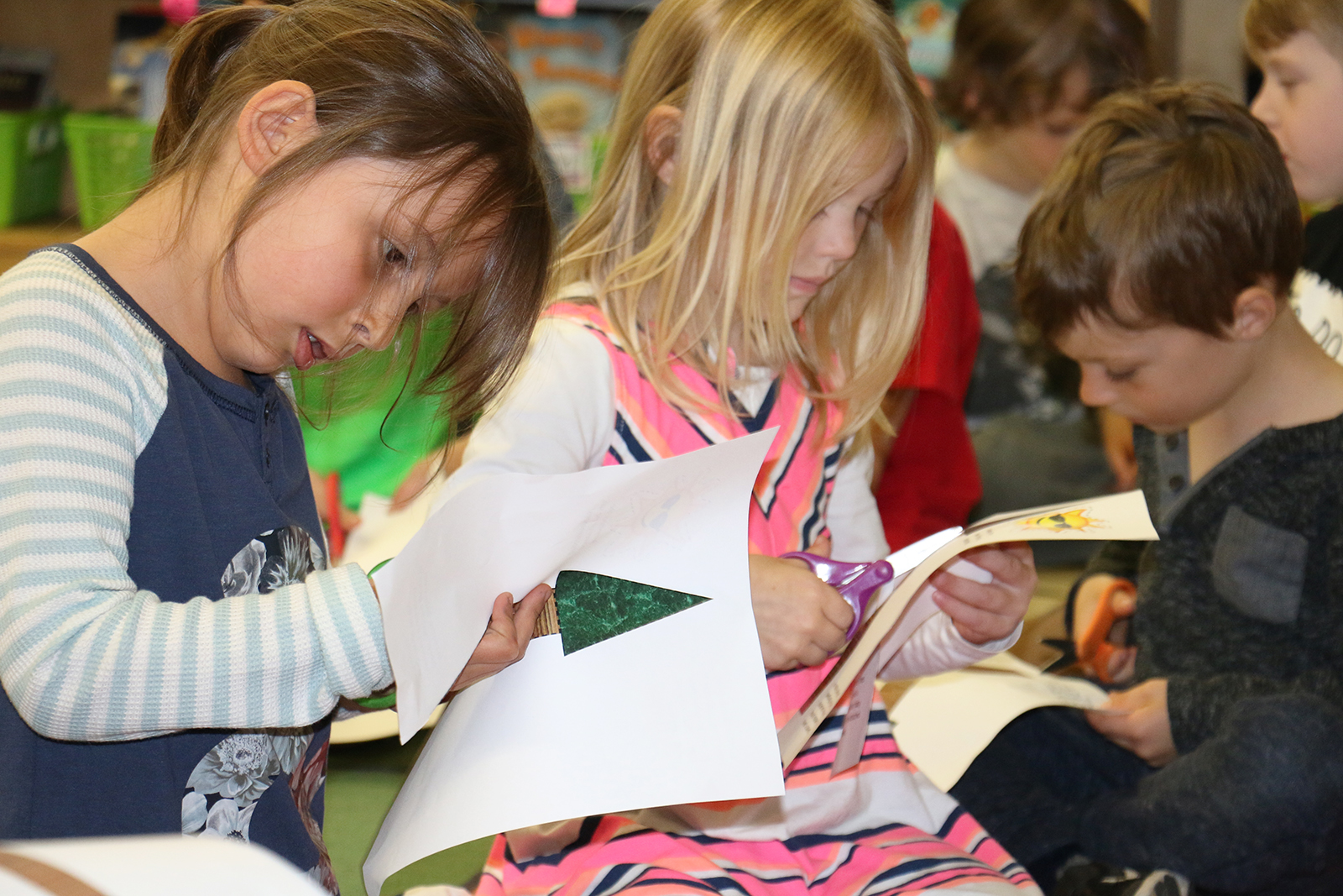 Three young children sit together in a classroom, reading books and holding paper cutouts.