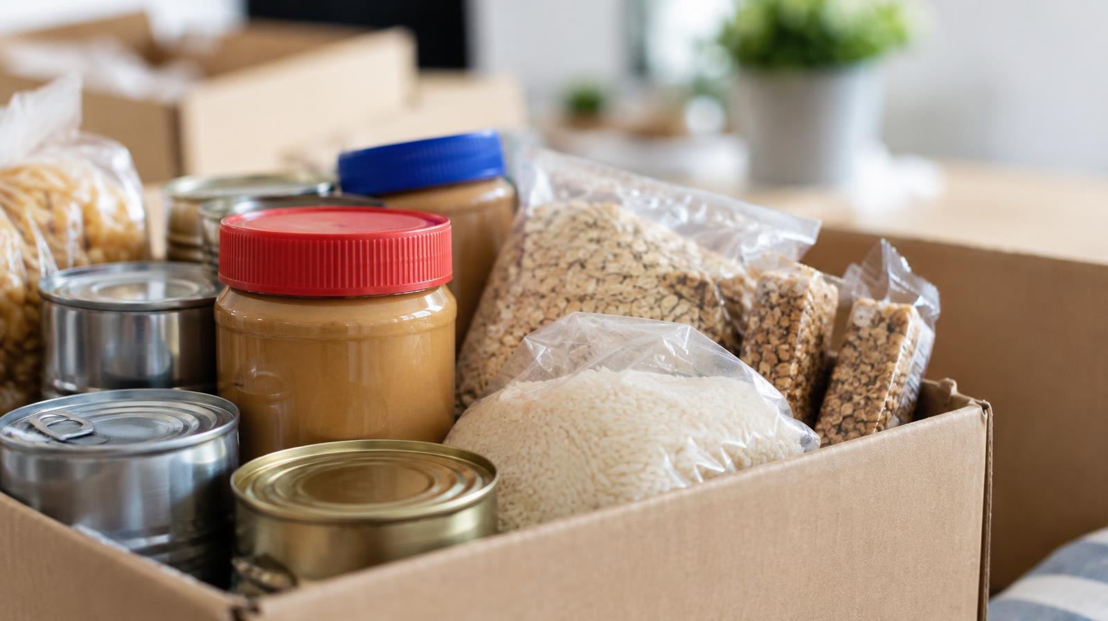 A cardboard box filled with canned goods, peanut butter jars, and packaged food items.