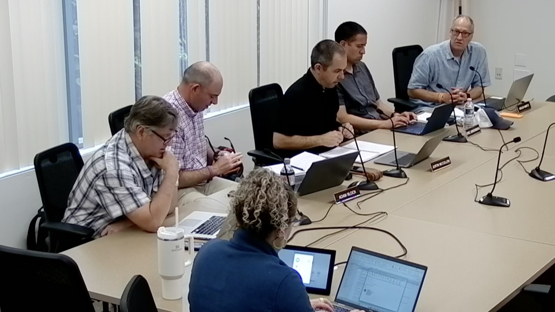 Six people seated around a conference table with laptops and name placards in a government meeting room.