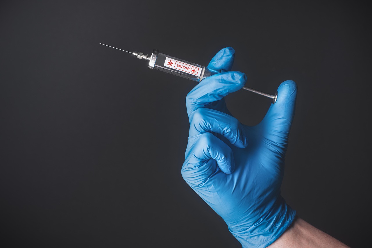 A gloved hand in a blue medical glove holds a syringe labeled "vaccine" against a dark background.