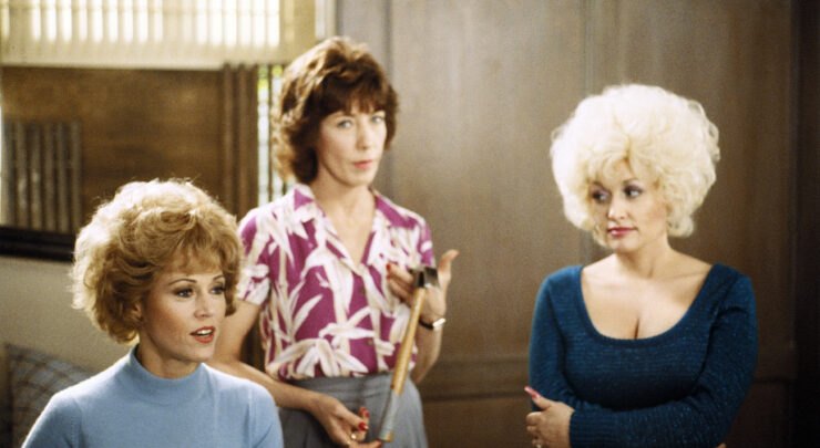 Three women in 1980s clothing stand and sit in an indoor setting in a film still.