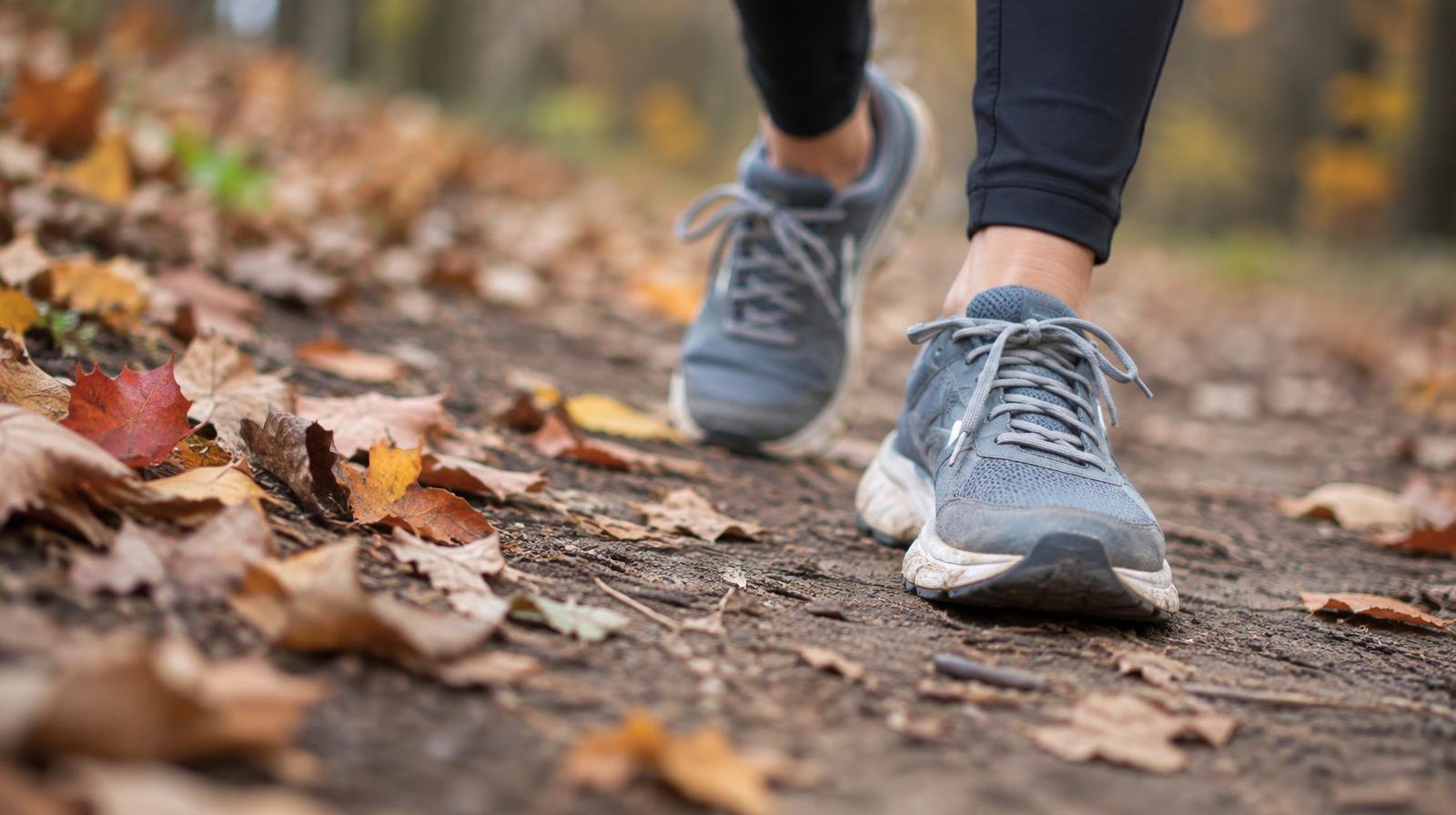 A pair of running shoes on a dirt trail surrounded by fallen autumn leaves.
