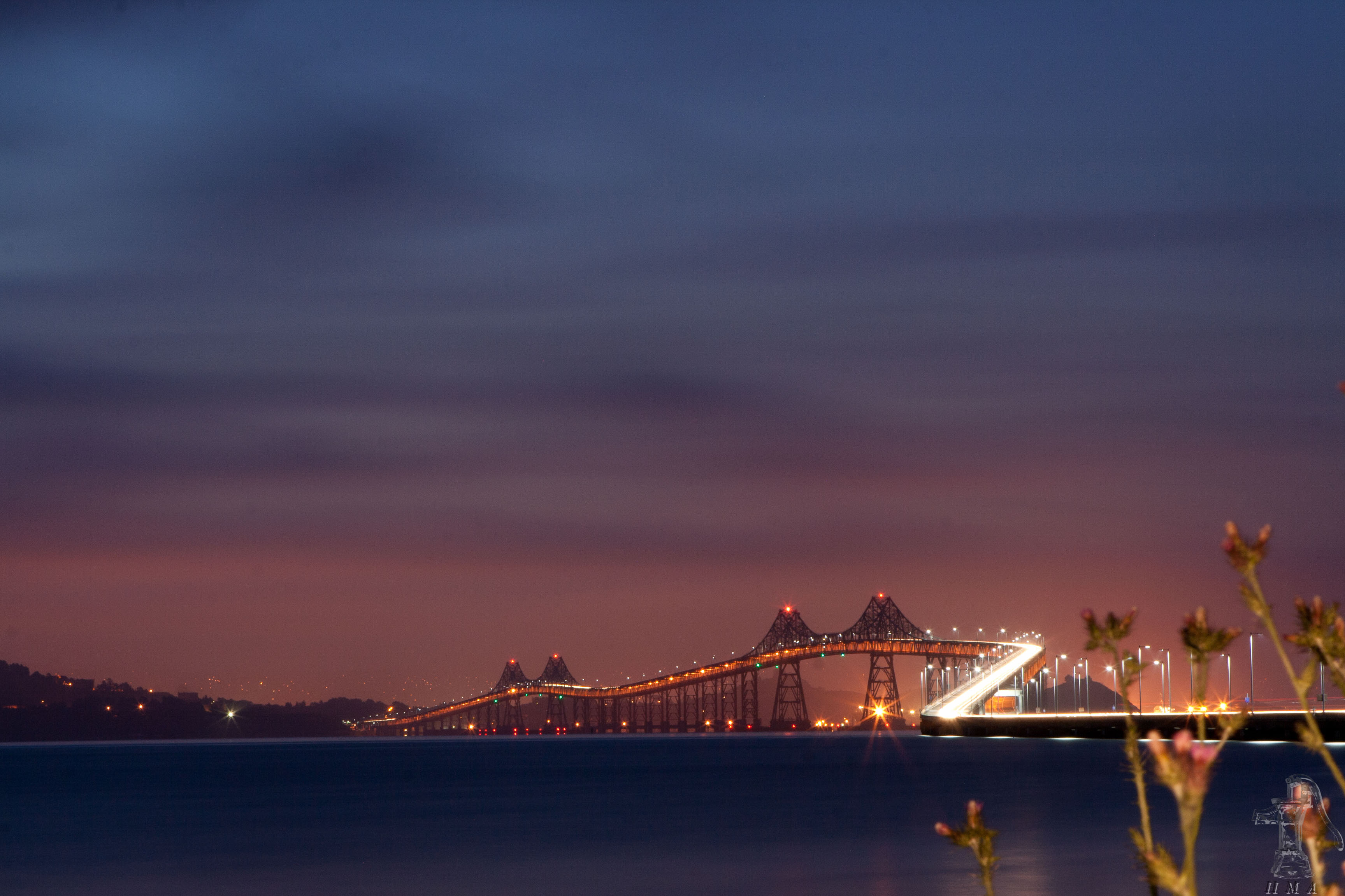 Bridge lit up at night with lights reflecting on calm bay water.
