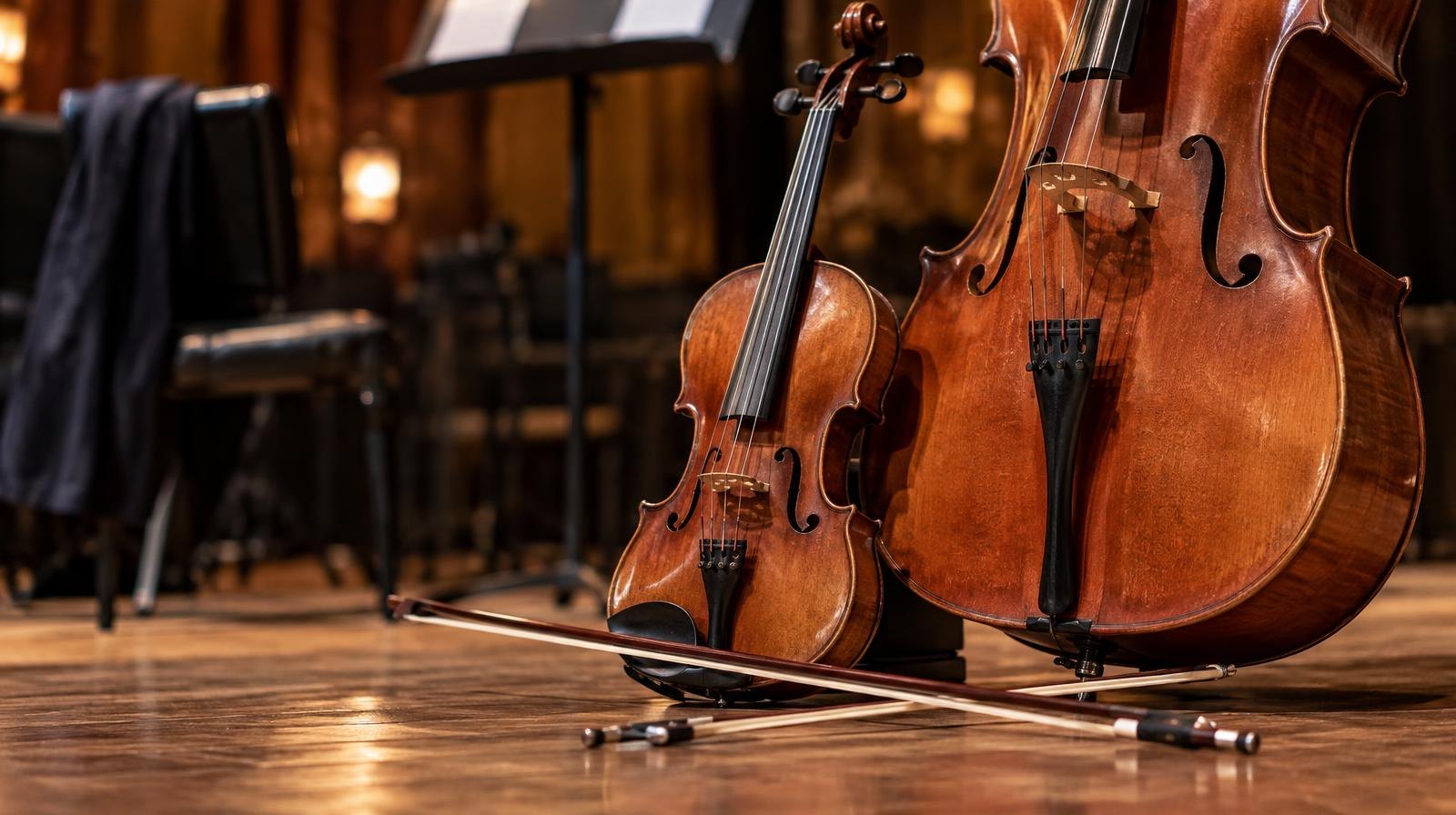 A fiddle and cello resting side by side on a wooden stage floor with crossed bows.
