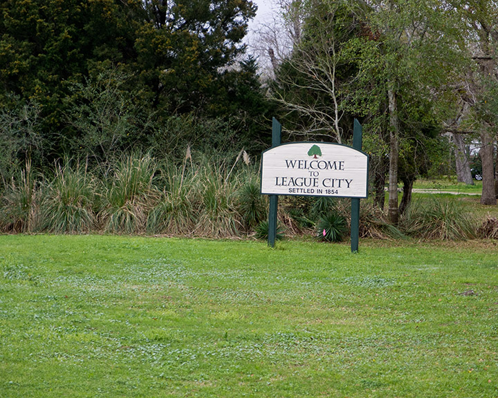 Green and white welcome sign reading 'Welcome to League City' on a grassy roadside.