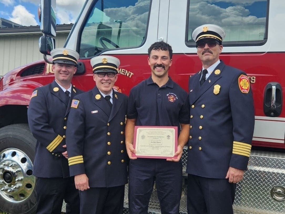Four people in fire department uniforms stand before a red fire truck, one holding a framed certificate.
