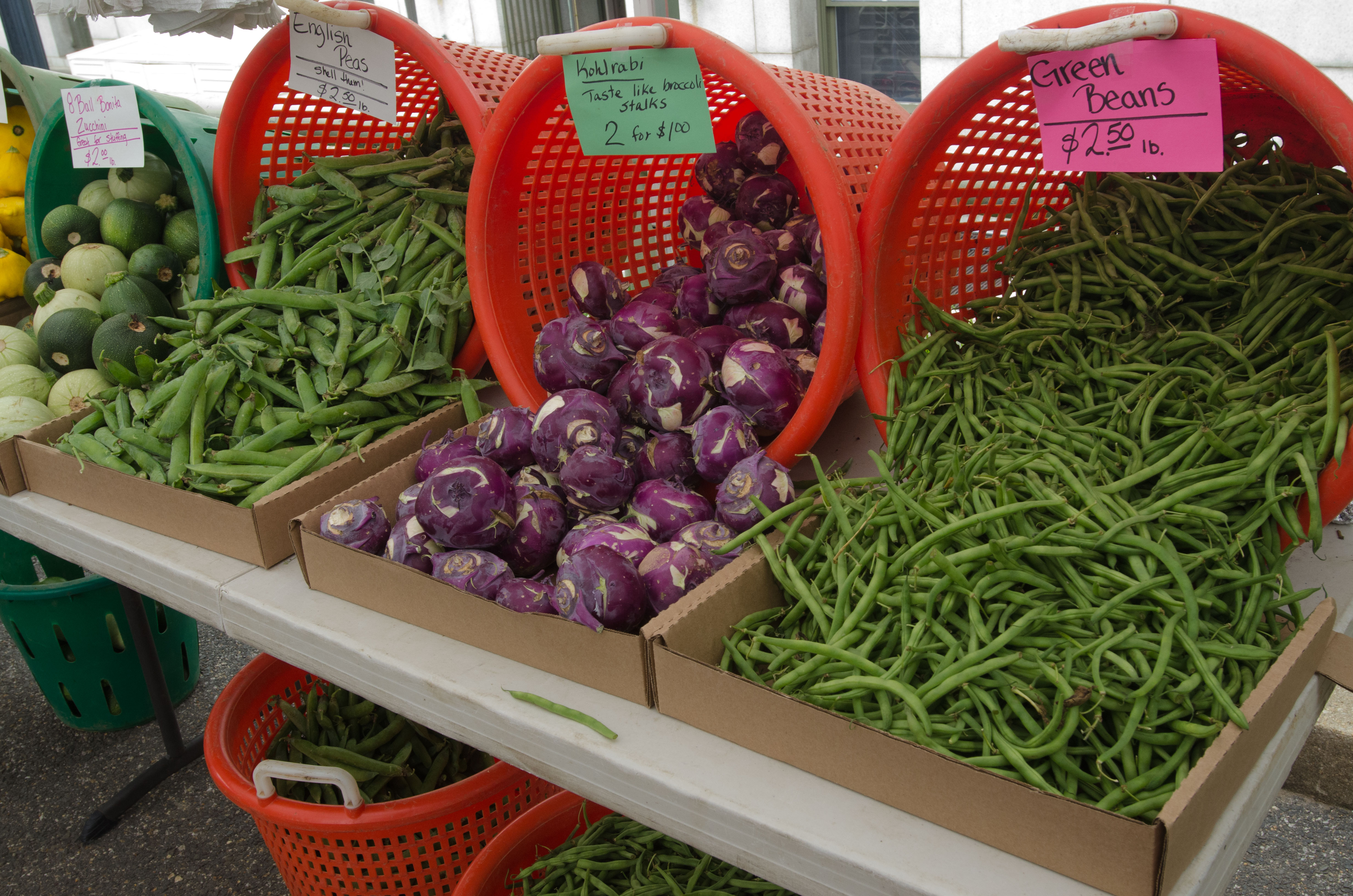 Fresh vegetables in red baskets and cardboard boxes at an outdoor farm stand with handwritten price signs.