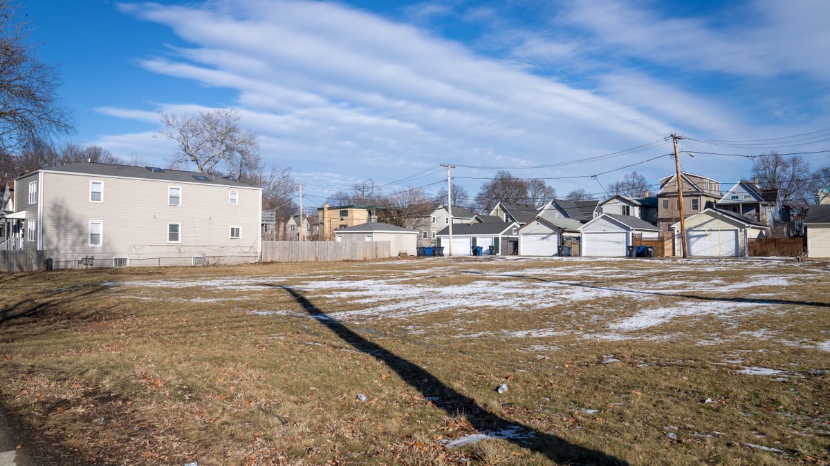 A snow-patched vacant lot surrounded by residential homes, garages, and a wooden fence under a cloudy sky.