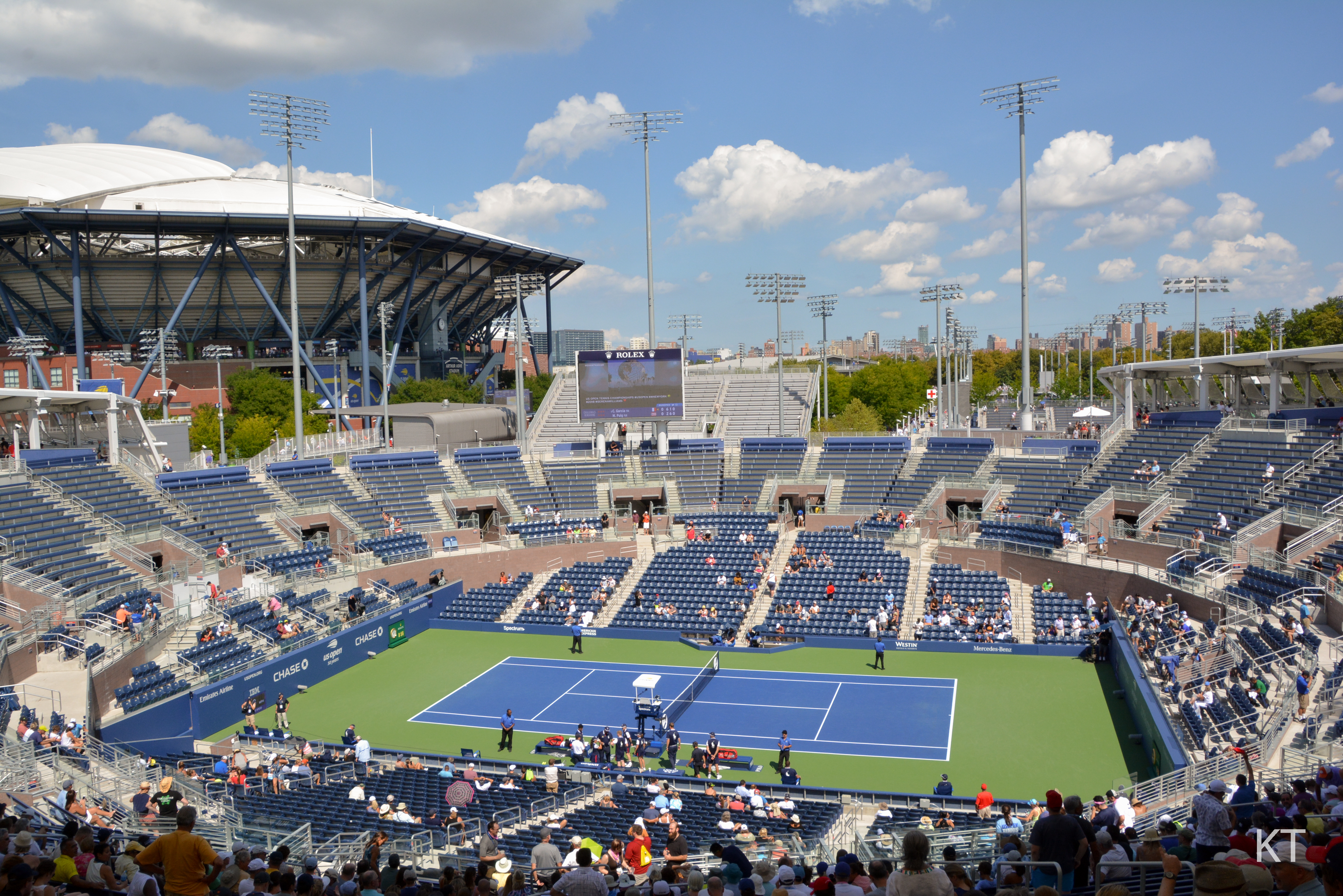 Aerial view of a blue outdoor tennis court surrounded by stadium seating under a partly cloudy sky.