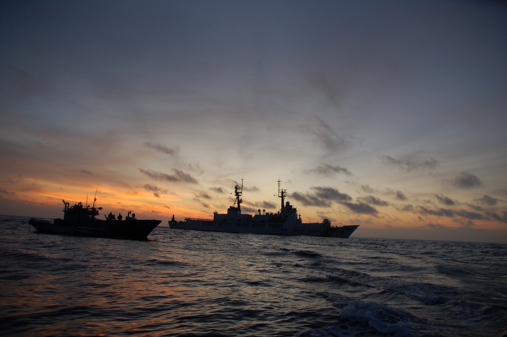 Large Coast Guard cutter and small open boat silhouetted together on open ocean at twilight.