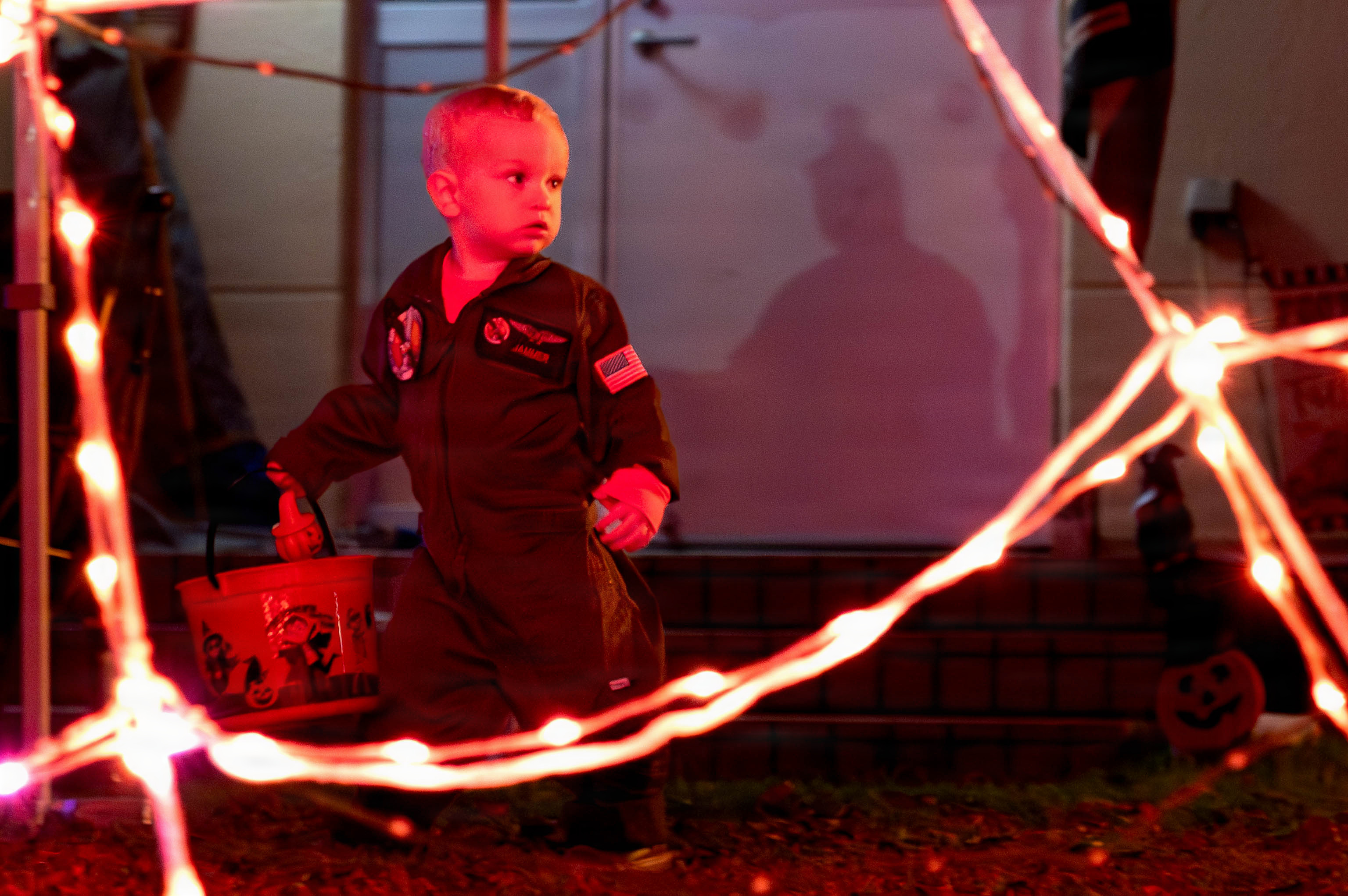 Young child in a costume holds a candy bucket surrounded by orange string lights at night.