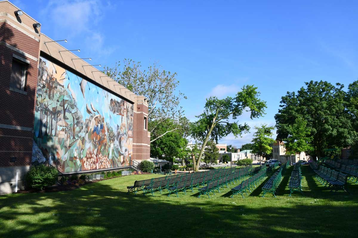 Outdoor concert venue with rows of green chairs on a lawn beside a brick building with a colorful mural.
