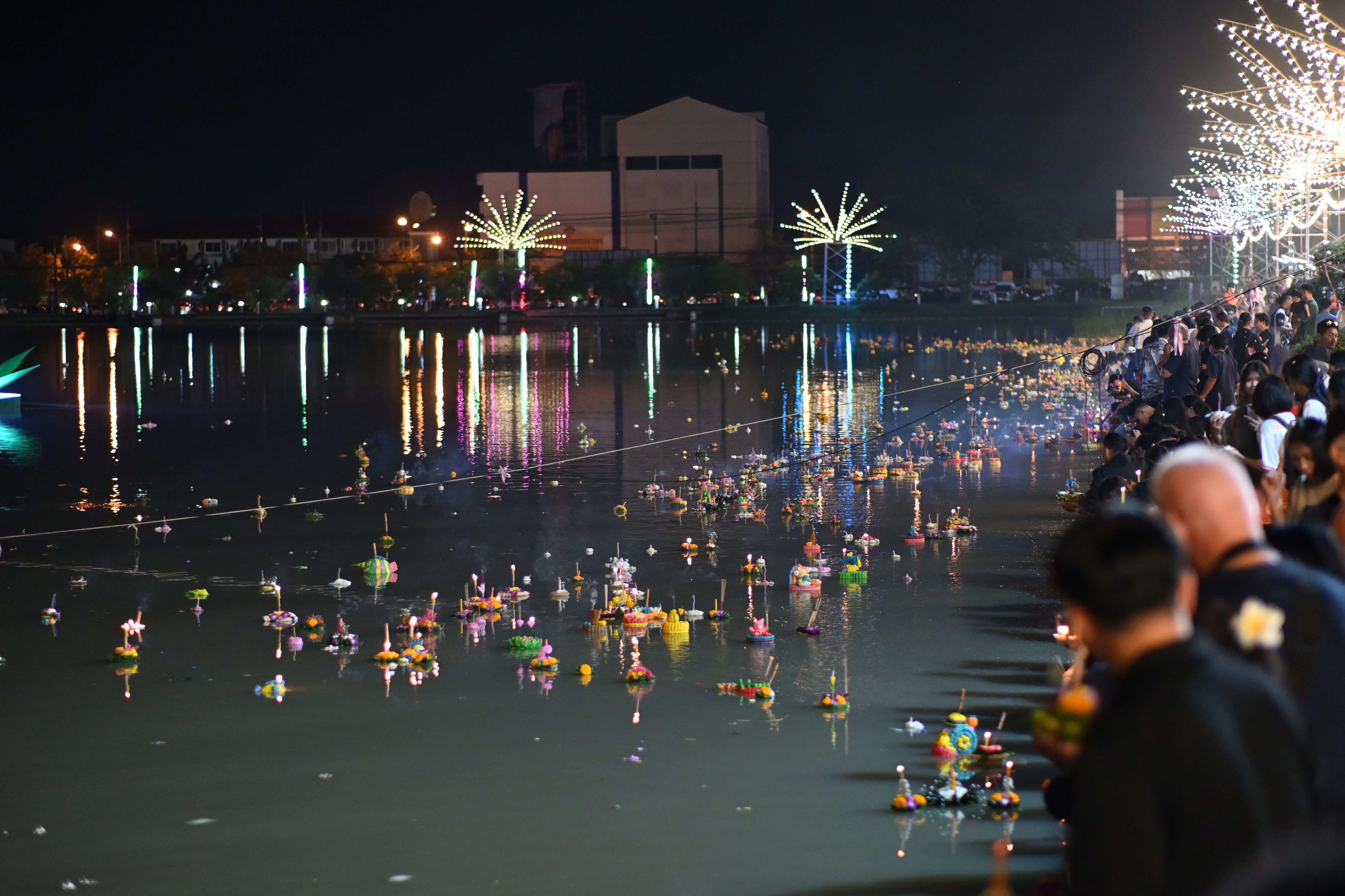Dozens of small glowing lanterns float on dark water at night, watched by crowds on shore.