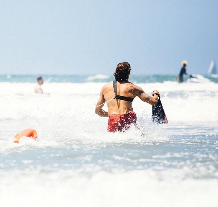 A lifeguard in red trunks runs into breaking ocean surf carrying rescue equipment.