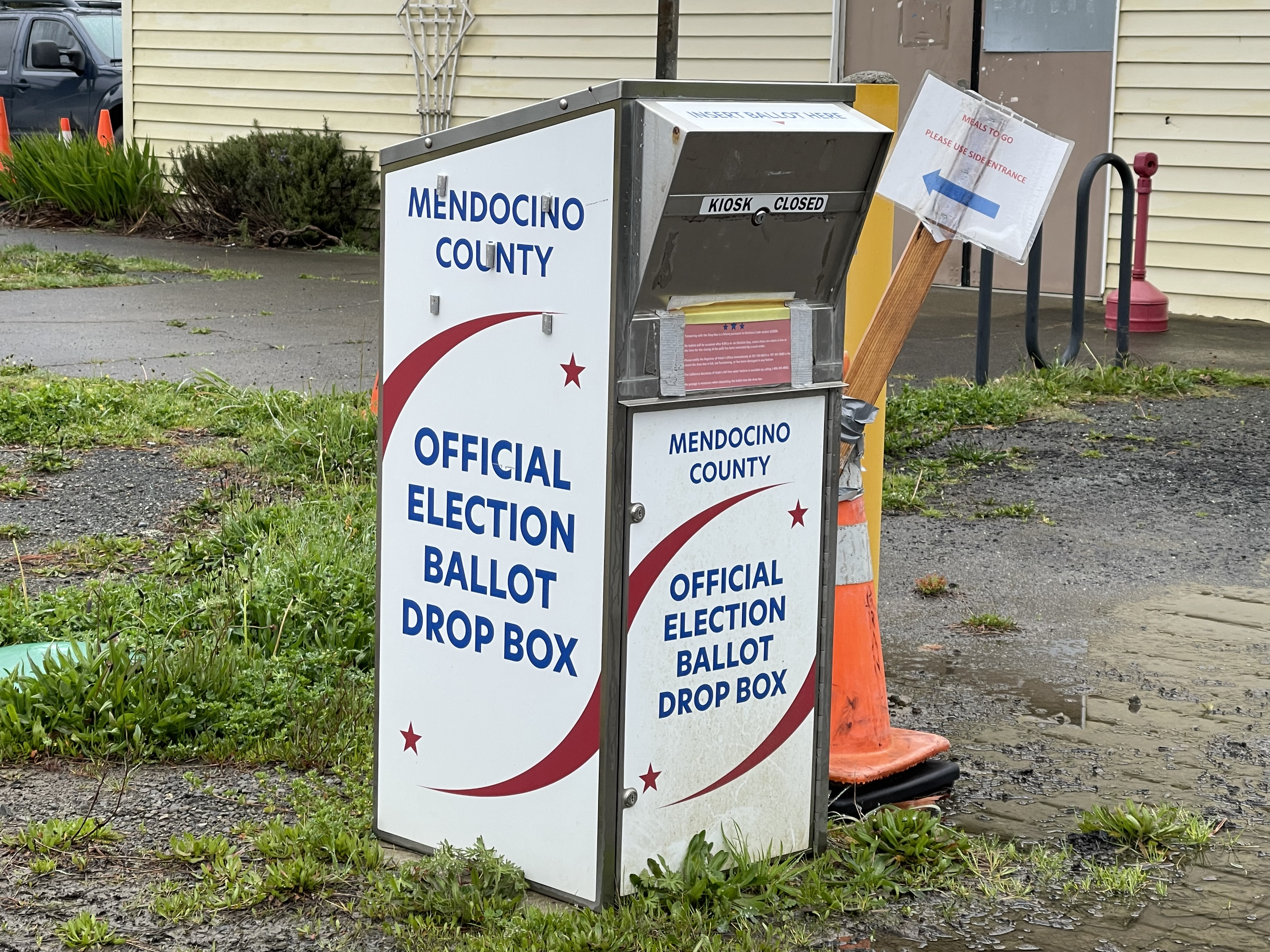 A white official election ballot drop box with red and blue graphics sits on a grassy area outdoors.