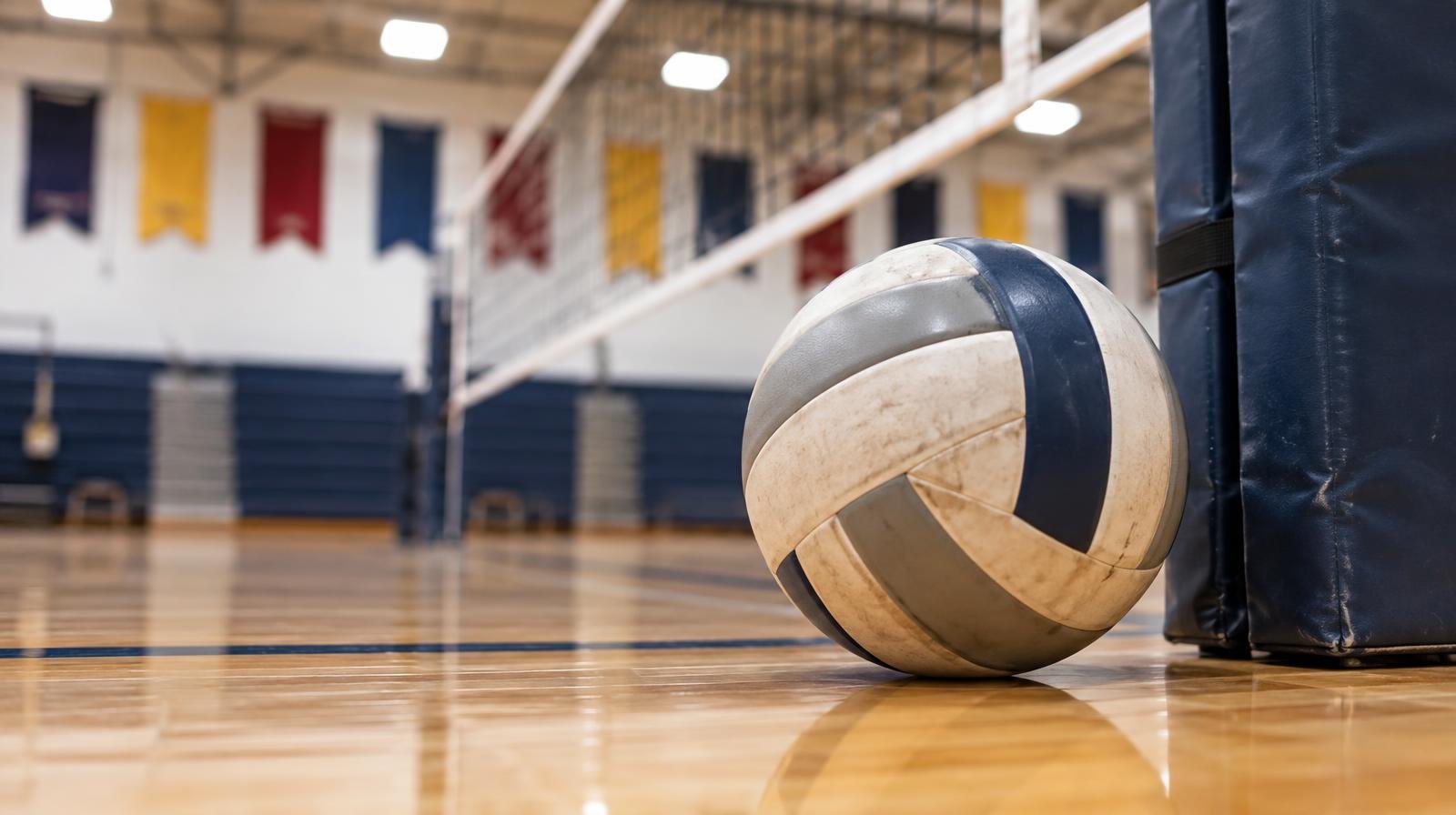 A volleyball resting on a hardwood gym floor next to a net post.