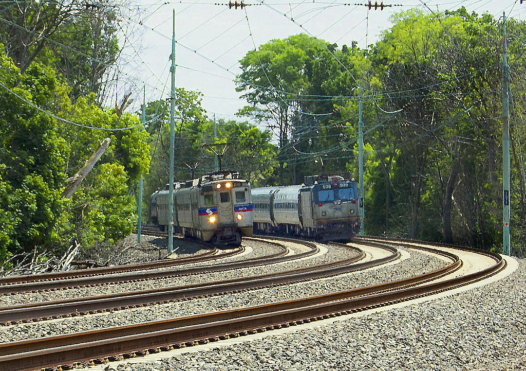 Two passenger trains passing on a curved four-track electrified rail corridor lined with trees