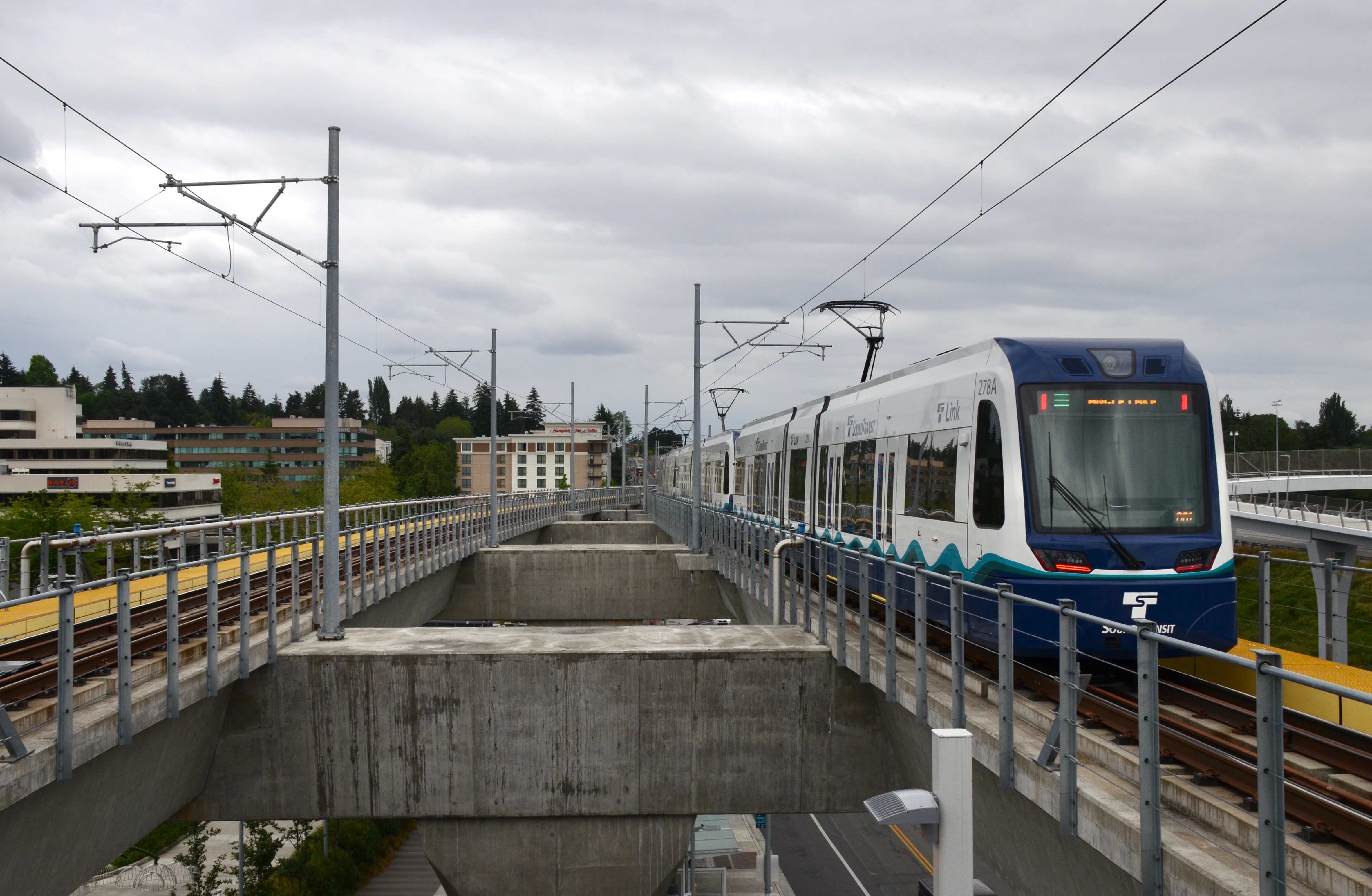 A light rail train pulling away from a station platform under an overcast sky.
