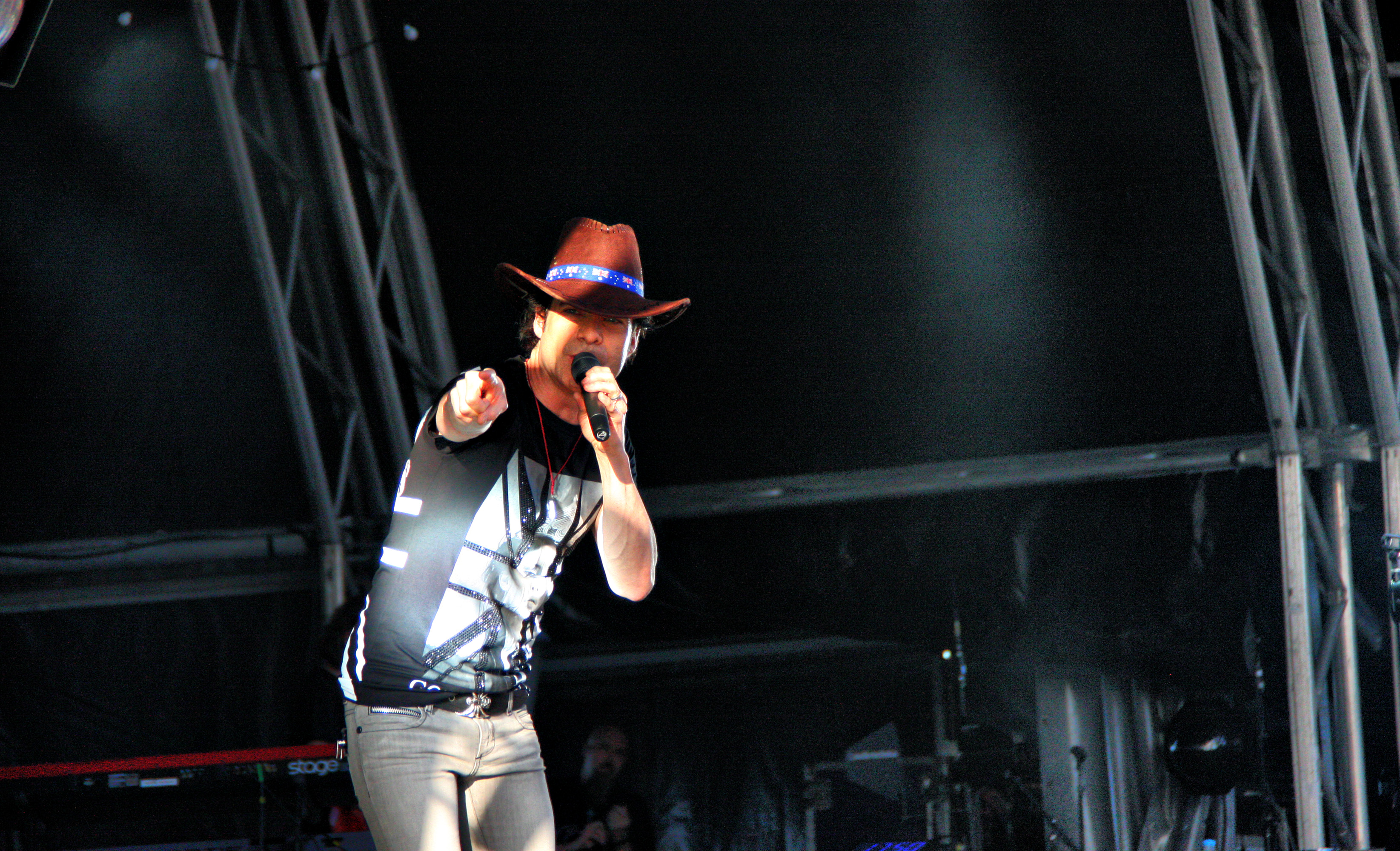 A performer in a red cowboy hat sings into a microphone and points toward the audience on a dark stage.
