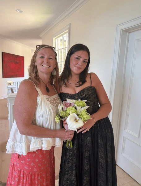 Two women standing together indoors, one holding a bouquet of flowers.