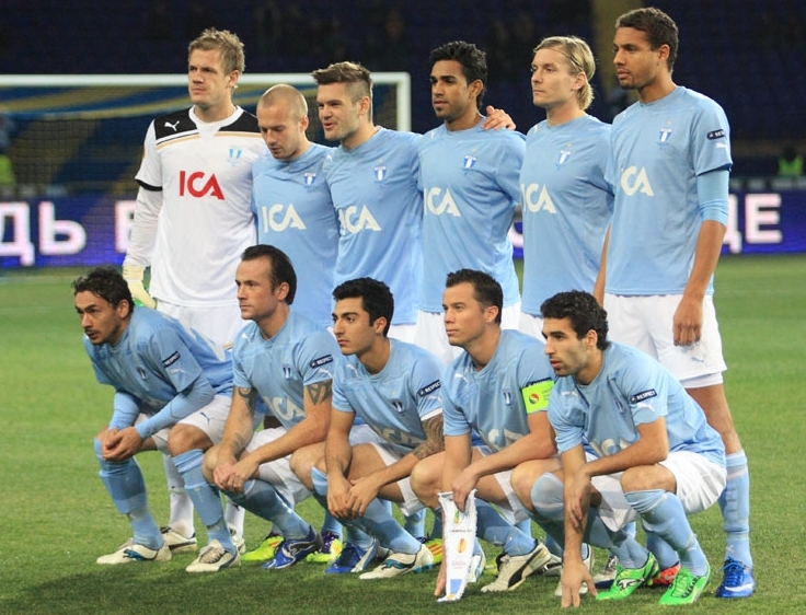 Football squad in light blue jerseys posed in two rows on a stadium pitch.