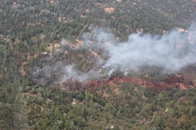 Aerial view of a wildfire burning through a forested hillside with smoke rising above the trees.