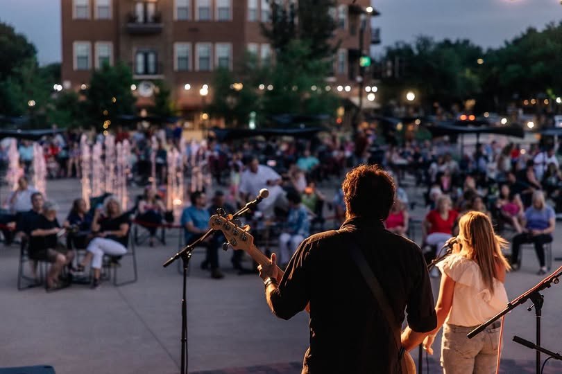 Guitarist performs on an outdoor stage facing a seated crowd at dusk, with illuminated fountains behind.