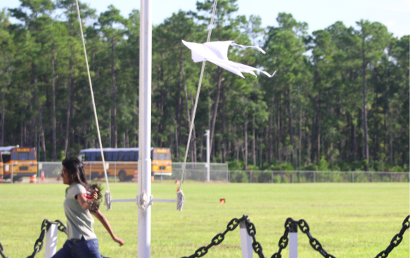 A white kite flies near a tall pole over a grassy school field.