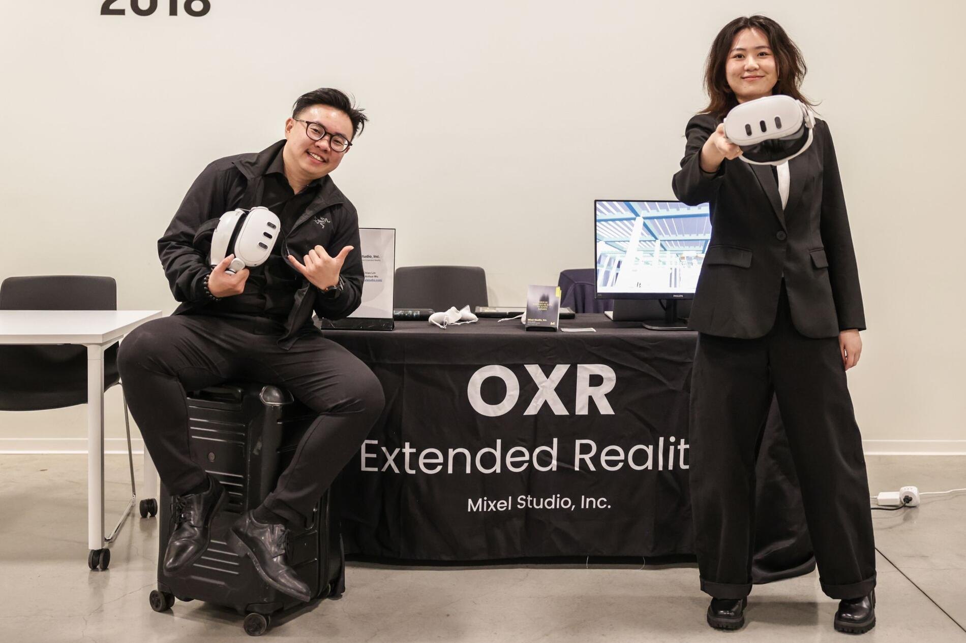 Two people smiling and holding white VR headsets in front of a branded OXR Extended Reality table.