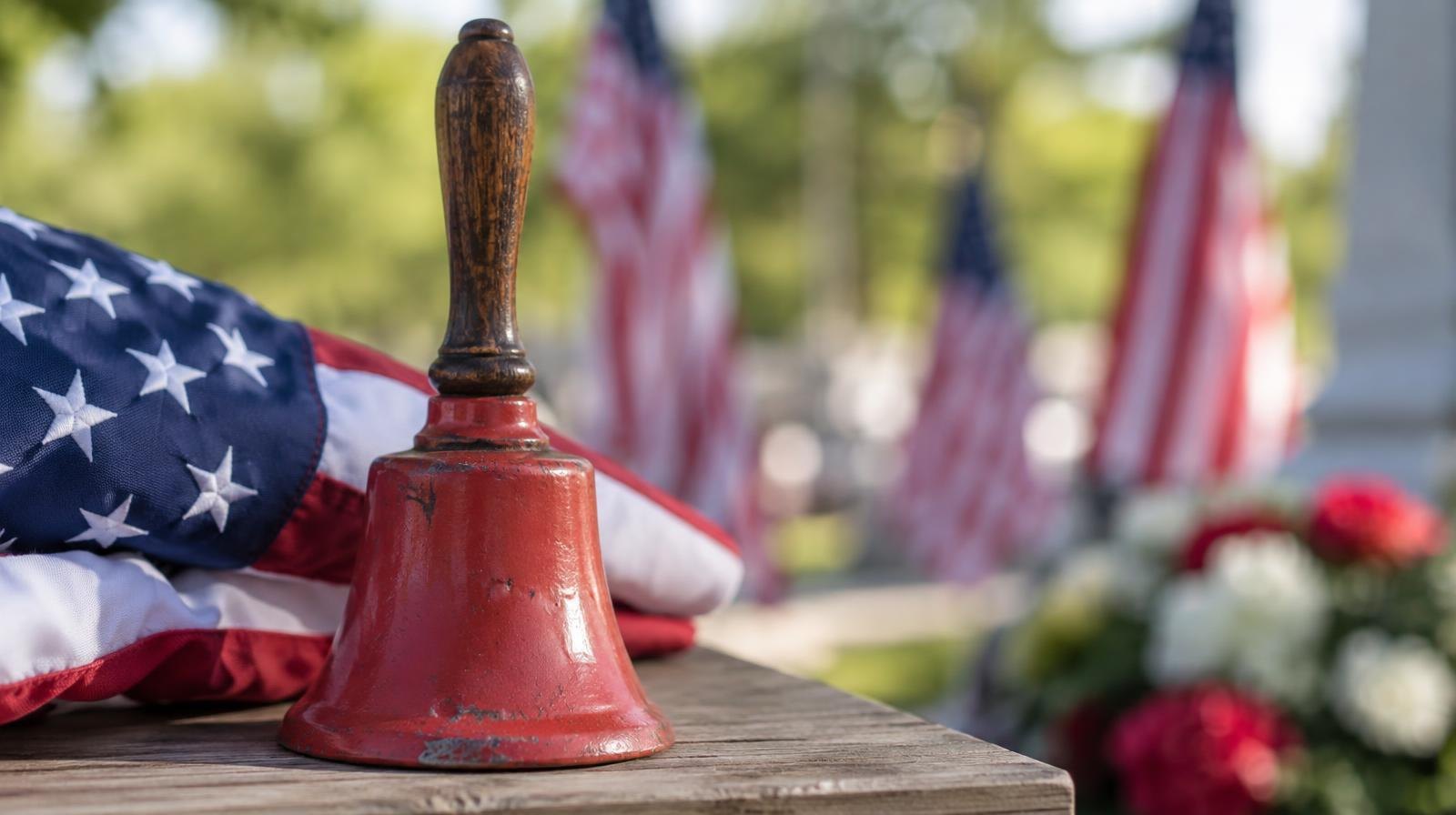 A red hand bell resting on a wooden surface next to a small American flag.