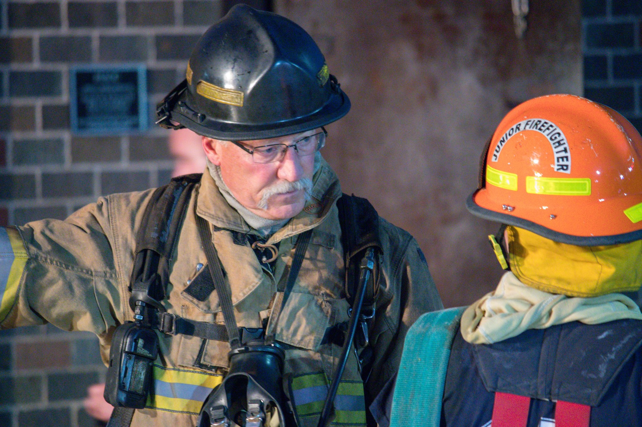 Senior firefighter in turnout gear speaks with a junior firefighter in an orange helmet at night.