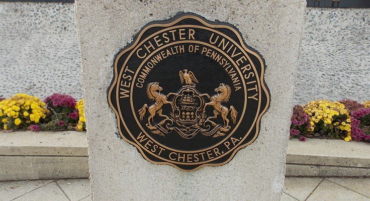 Bronze university seal mounted on a stone pillar, flanked by flowering plants.