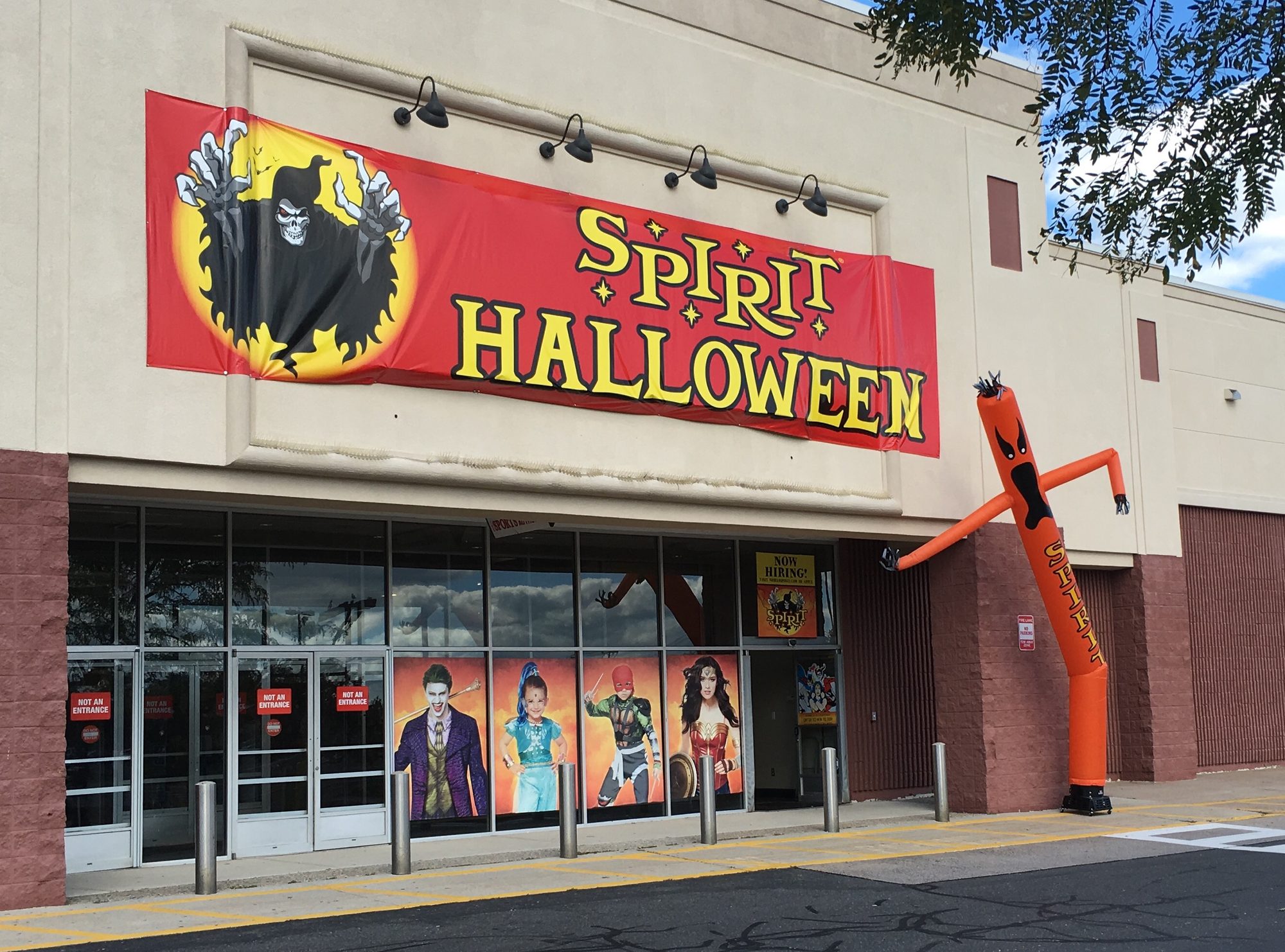 Exterior of a Spirit Halloween store with a red promotional banner over the entrance and an inflatable tube figure outside.