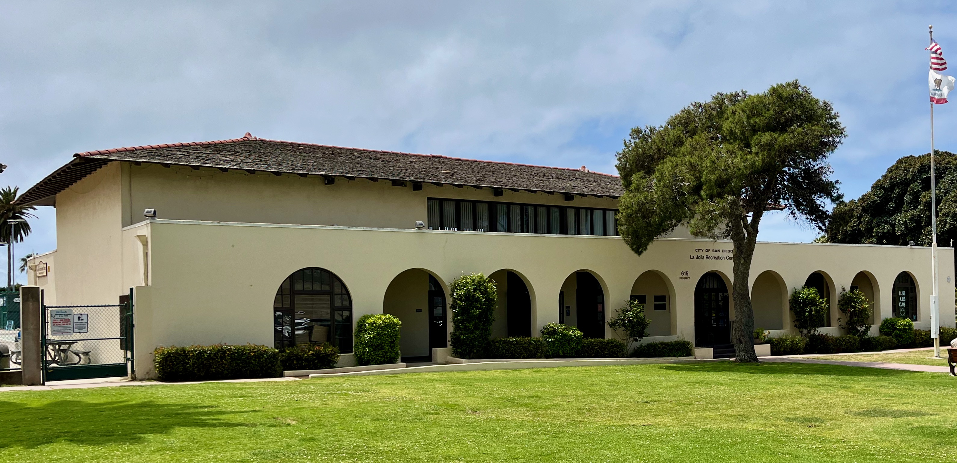 Single-story stucco building with arched colonnades, green lawn, and American flag on a pole.