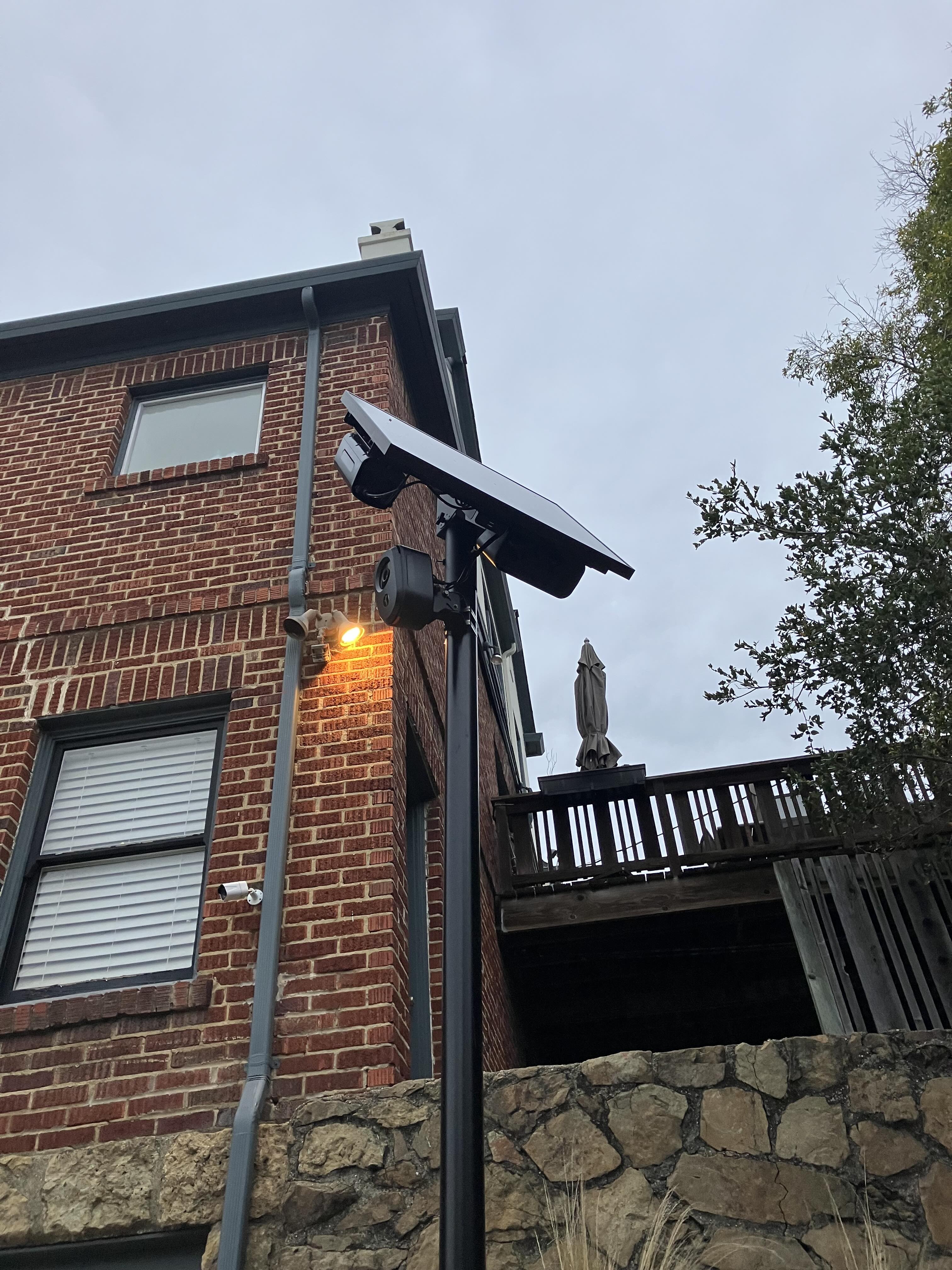 A Flock automated license plate reader camera on a pole with a solar panel in front of a brick house.