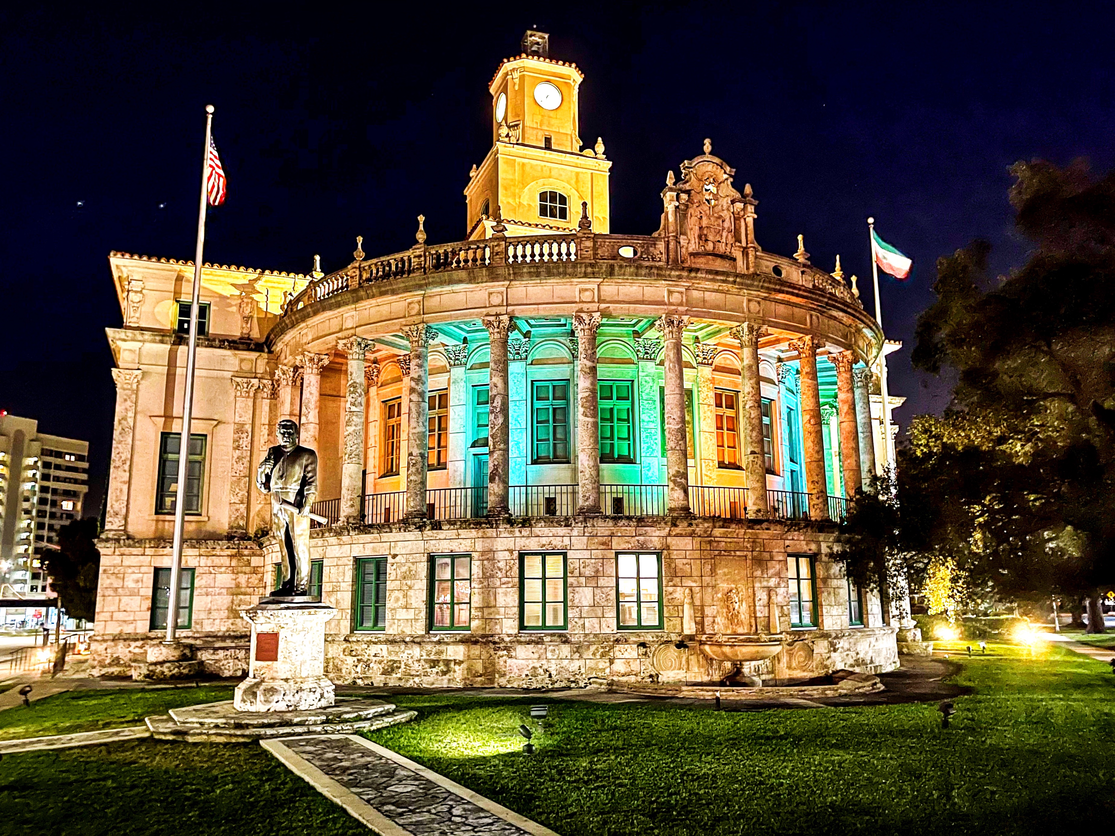 Neoclassical city hall building illuminated in teal and orange at night, with a statue in front.