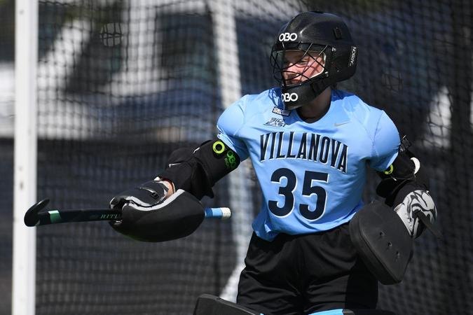 Field hockey goalkeeper in light blue jersey number 35 and full protective gear stands in front of a net.