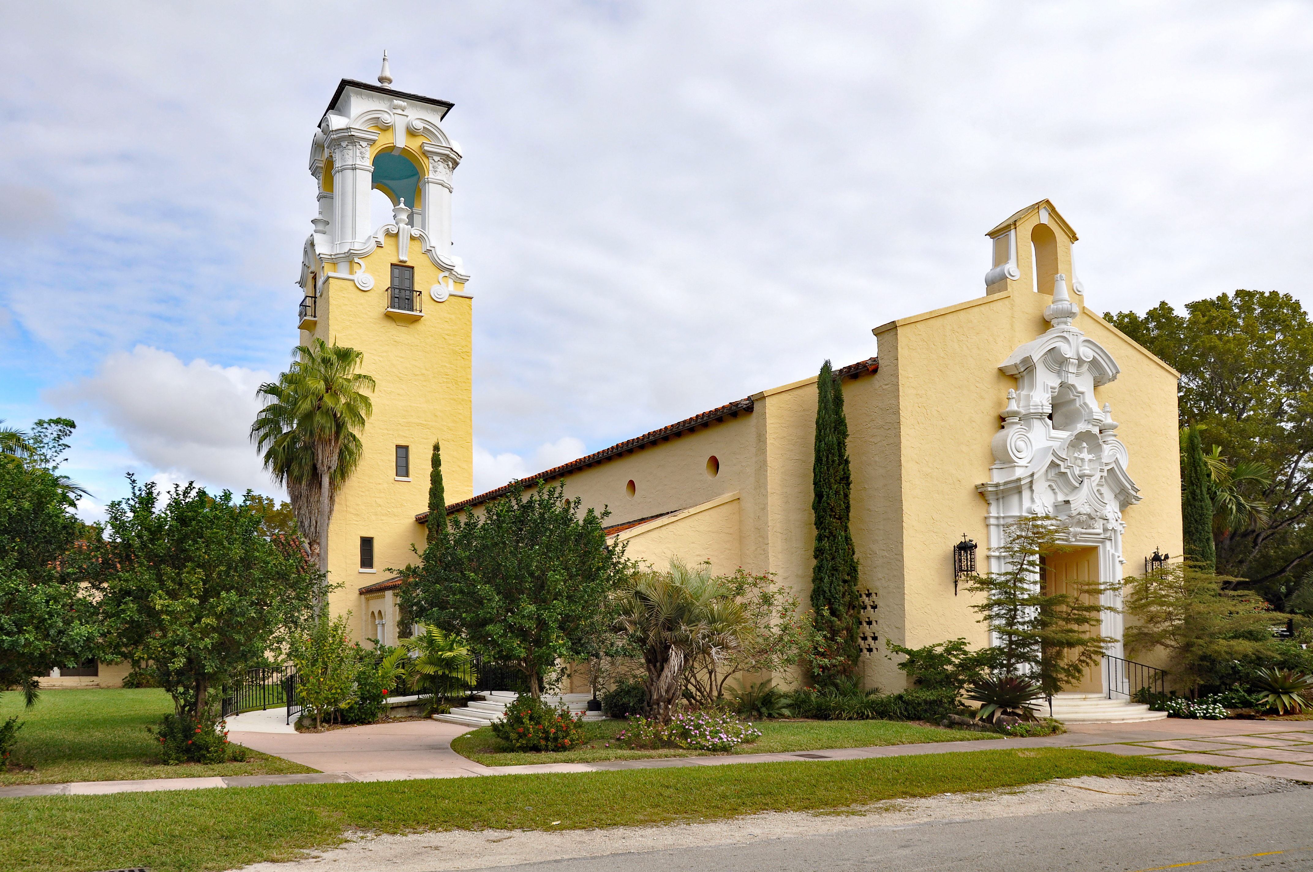 Side view of a pale yellow stucco church with a white bell tower and red tile roof.