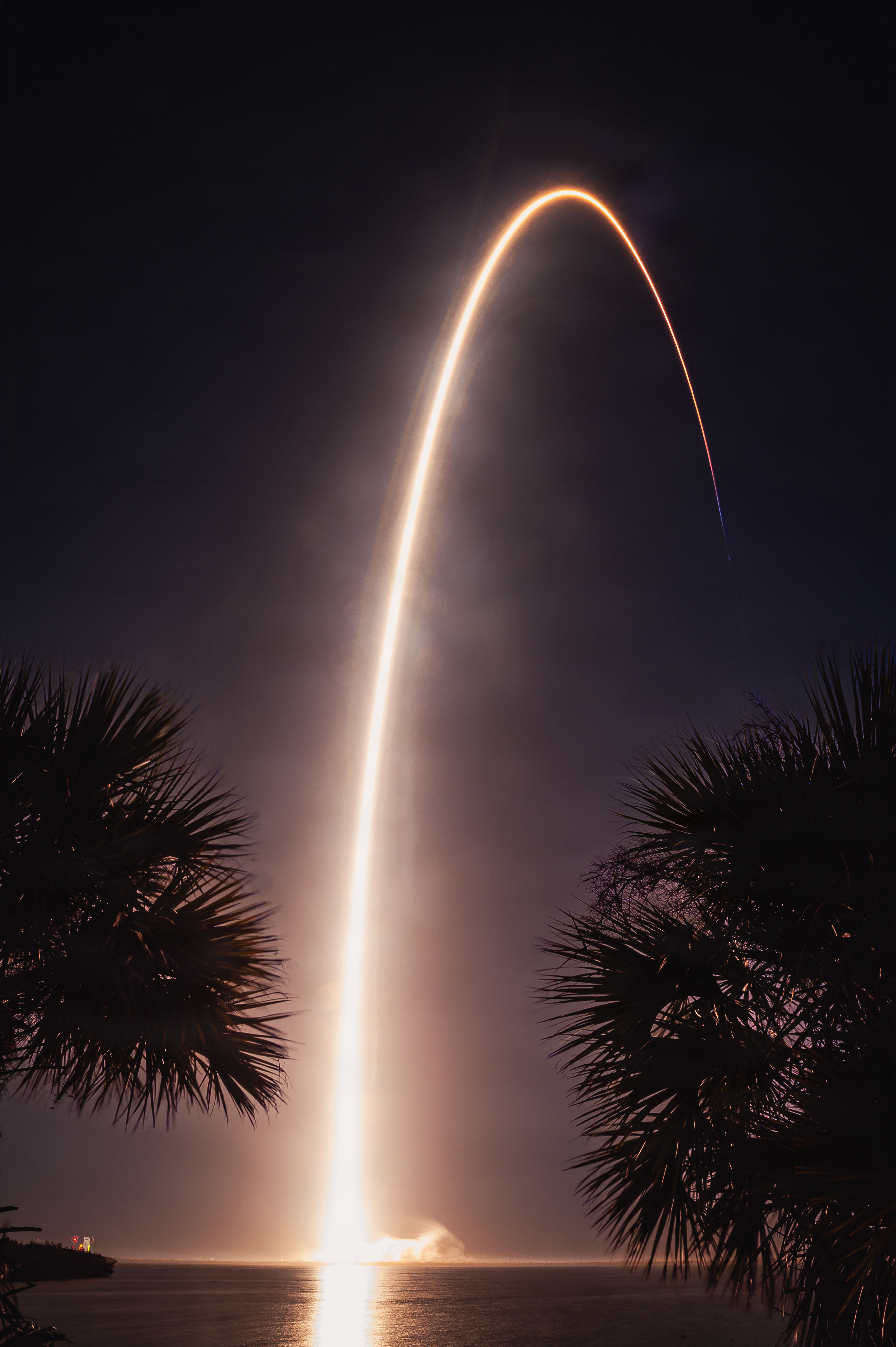 Long-exposure photo of a rocket's bright arc over water with palm trees silhouetted below.