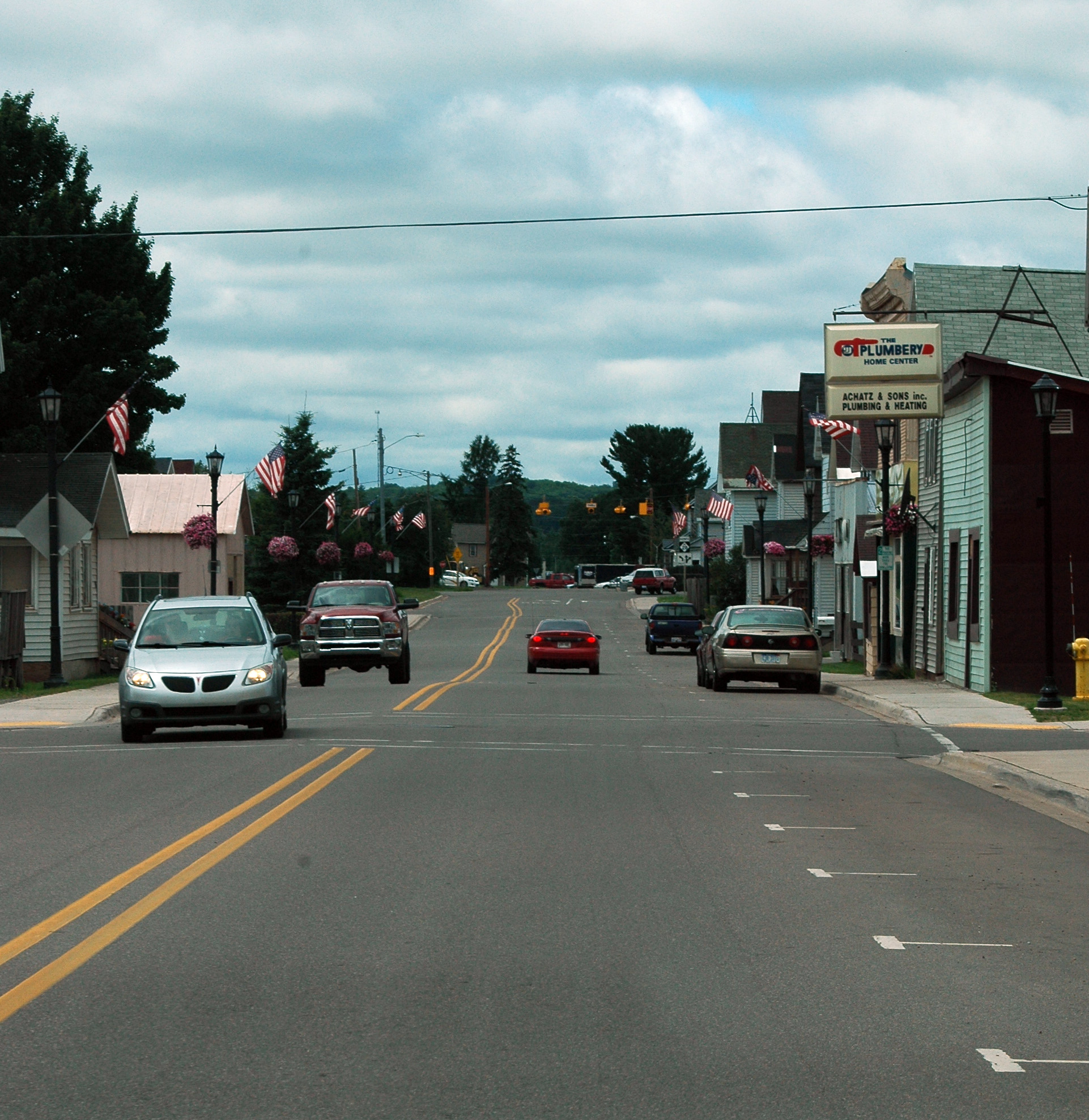 Small-town main street with storefronts, American flags on light poles, and passing cars.
