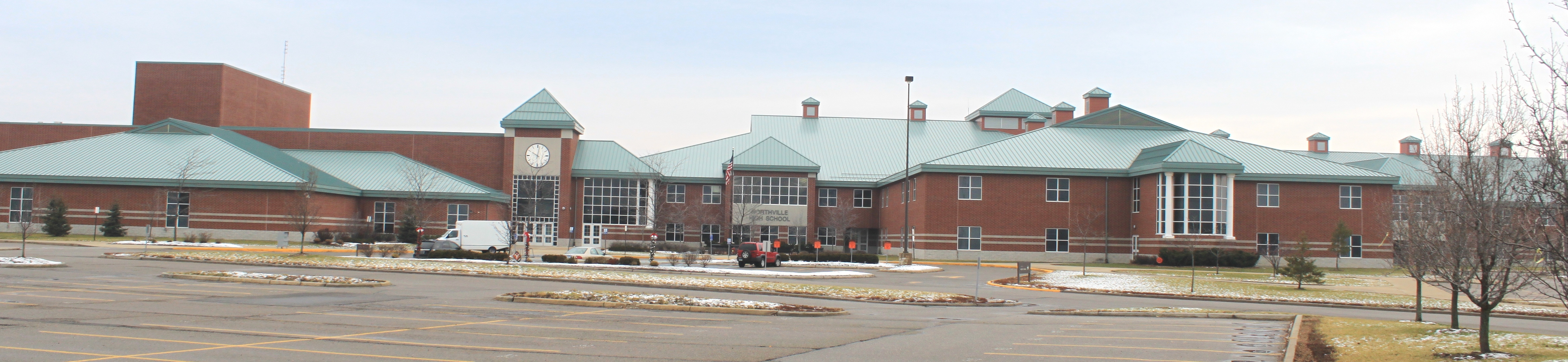 Panoramic view of a brick school building with a green roof and clock tower, parking lot in front.