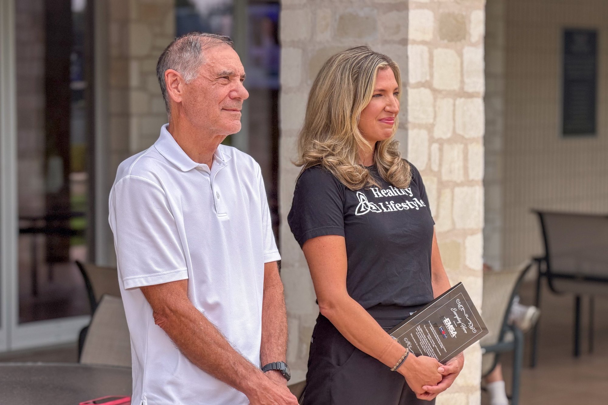 A man in a white polo and a woman in a black shirt stand together outdoors, the woman holding a dark award plaque.