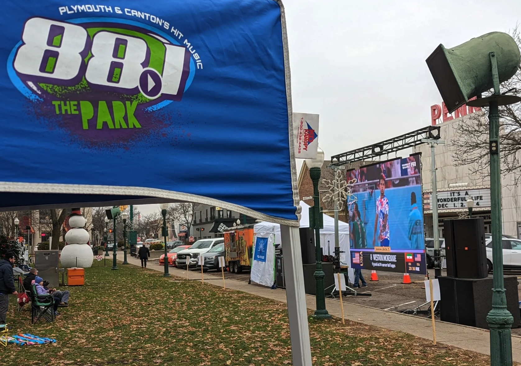 An 88.1 The Park banner flies above a downtown Plymouth community event, with a large outdoor screen showing a soccer broadcast in the background.