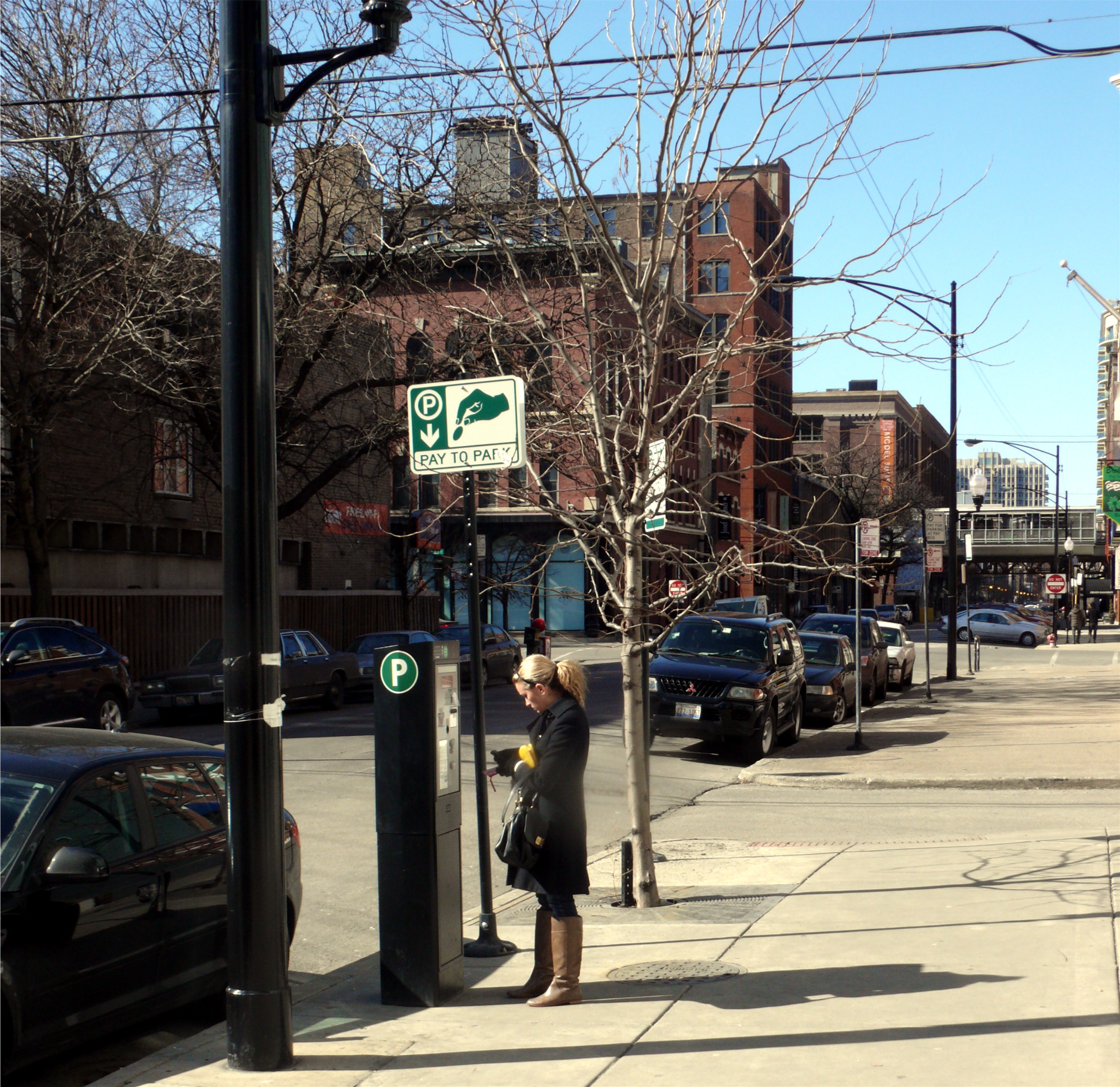 A woman uses a pay-to-park kiosk on a city sidewalk with parked cars nearby.