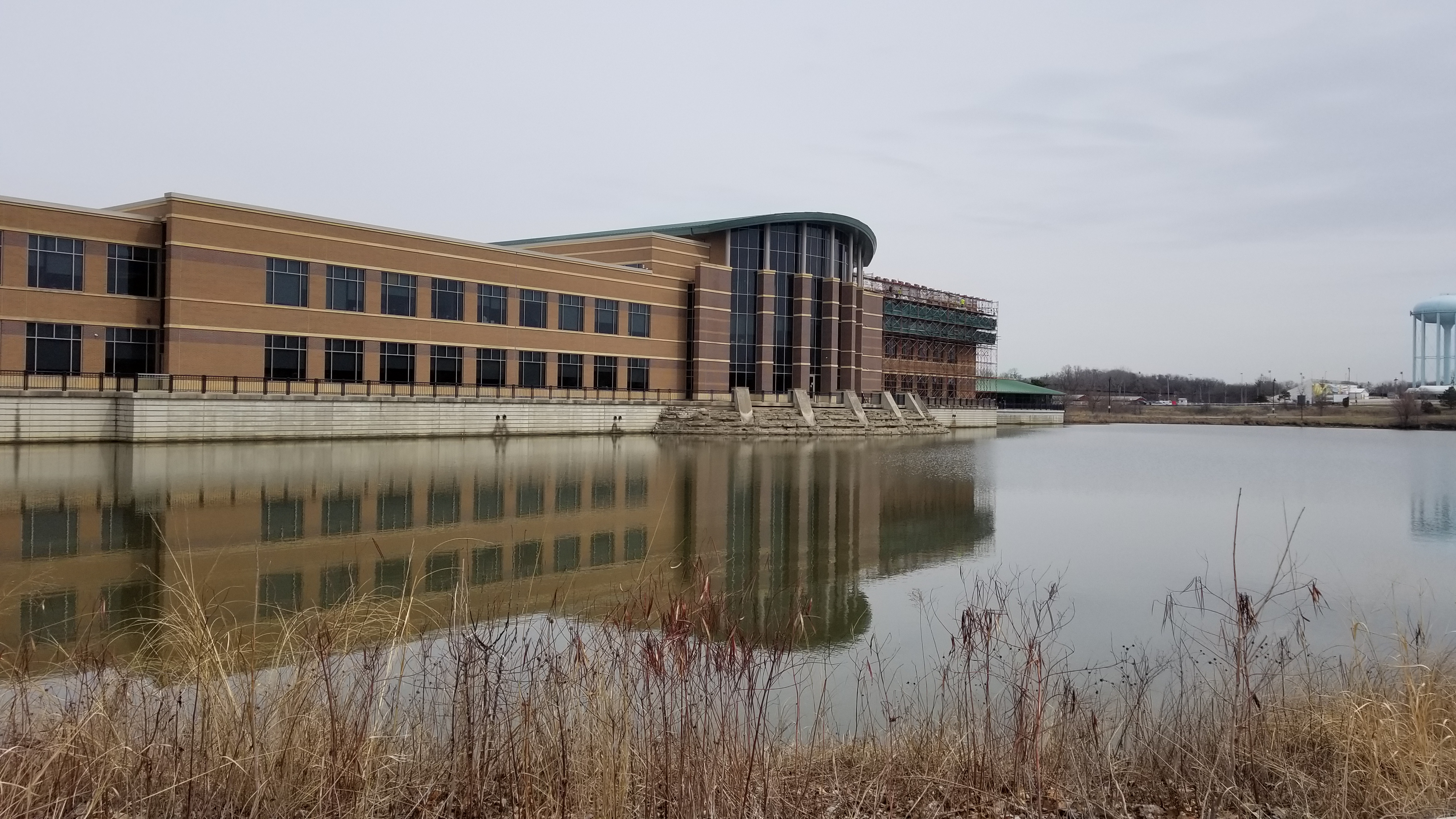 Multi-story brick government building reflected in a pond with bare winter vegetation in the foreground.