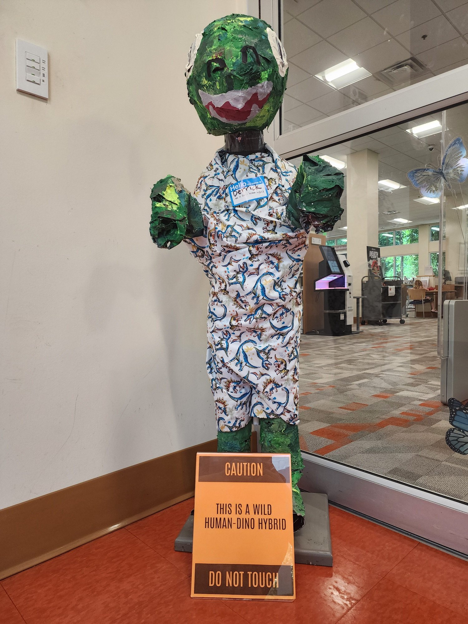 A green papier-mâché dinosaur figure wearing dinosaur-print pajamas stands in a library lobby next to a caution sign.