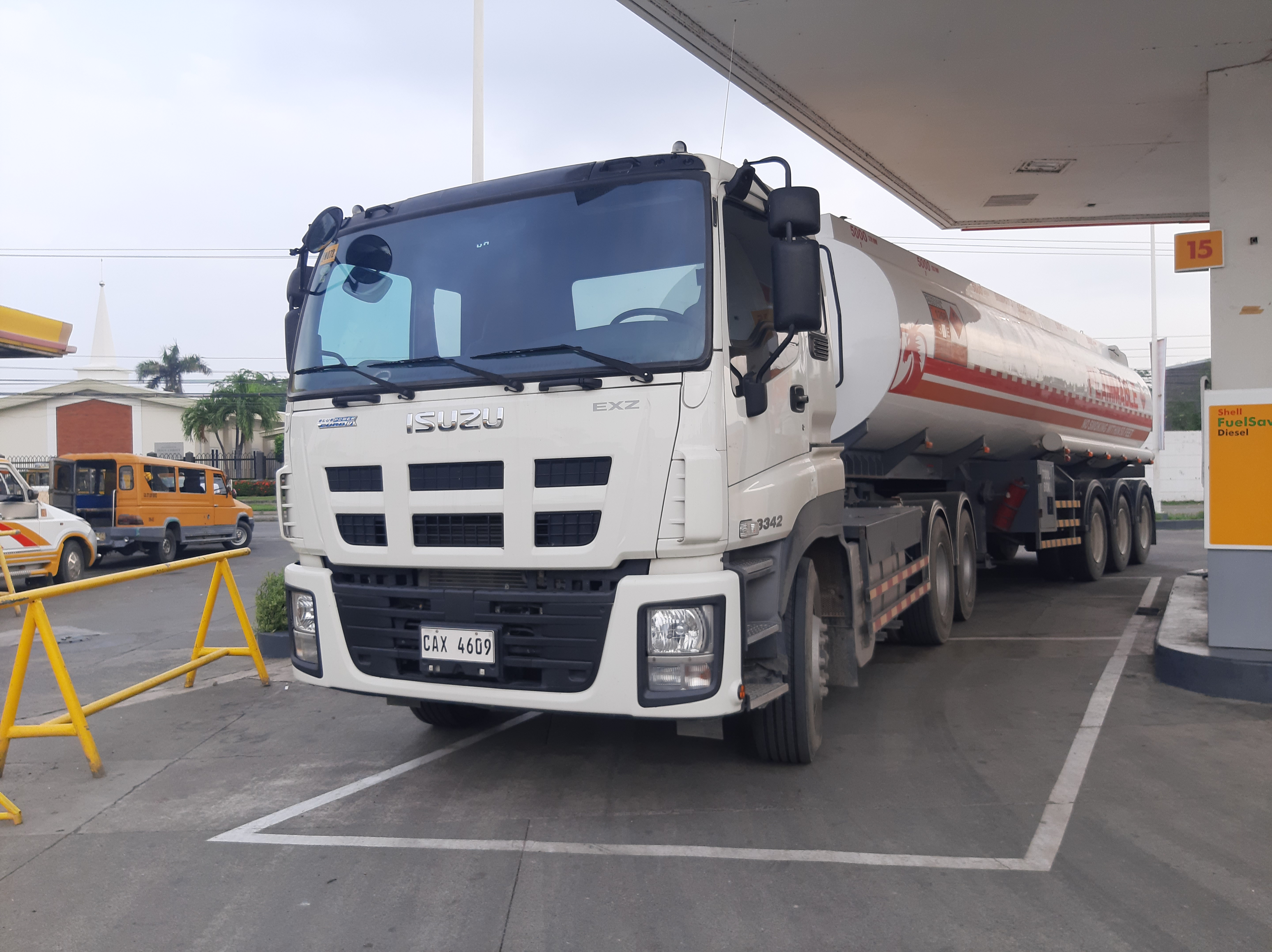 A white tractor-head truck with a red-and-white fuel tanker trailer parked at a gas station.