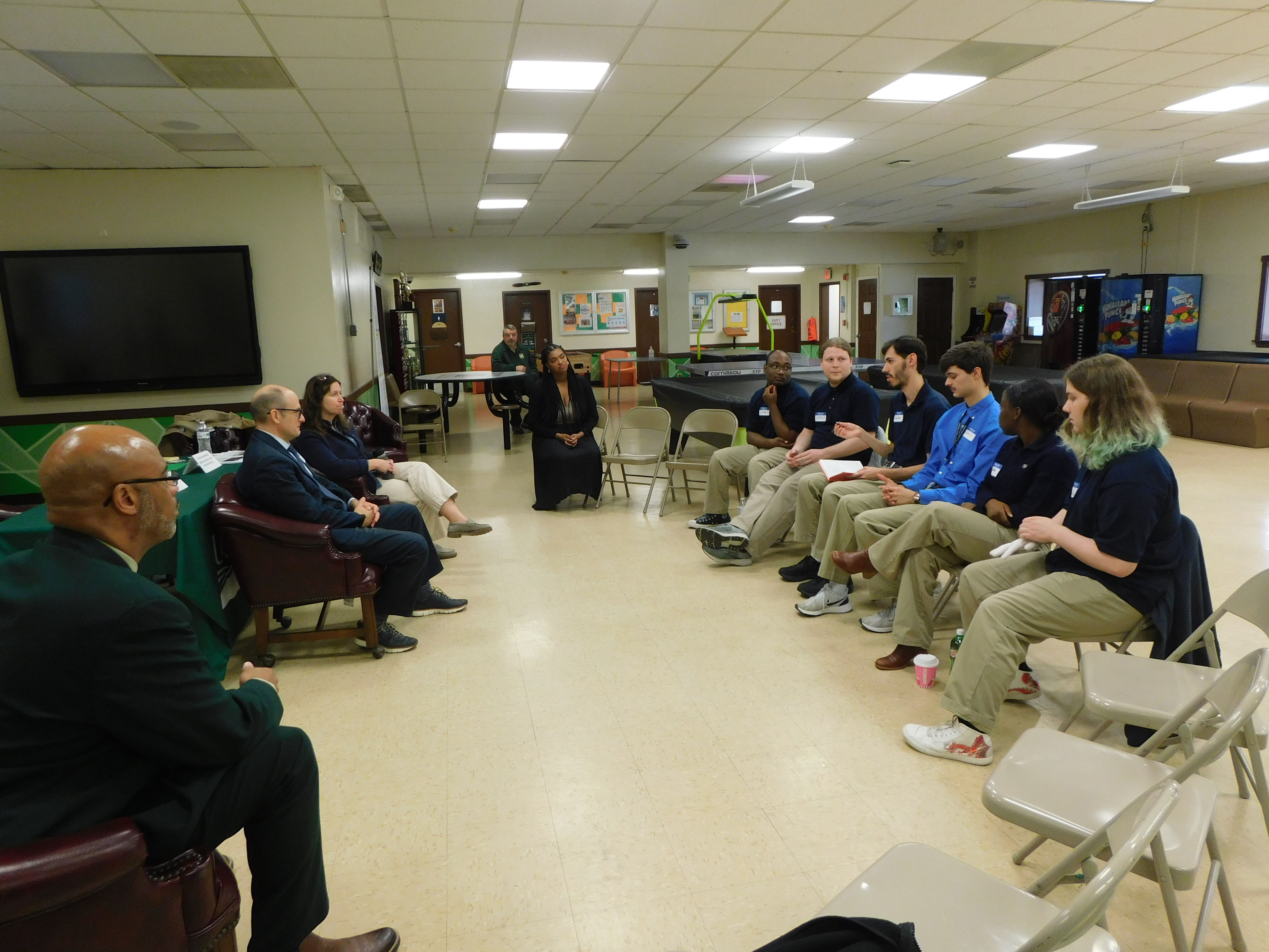 Young people in matching uniforms and adults in suits seated in a circle indoors.