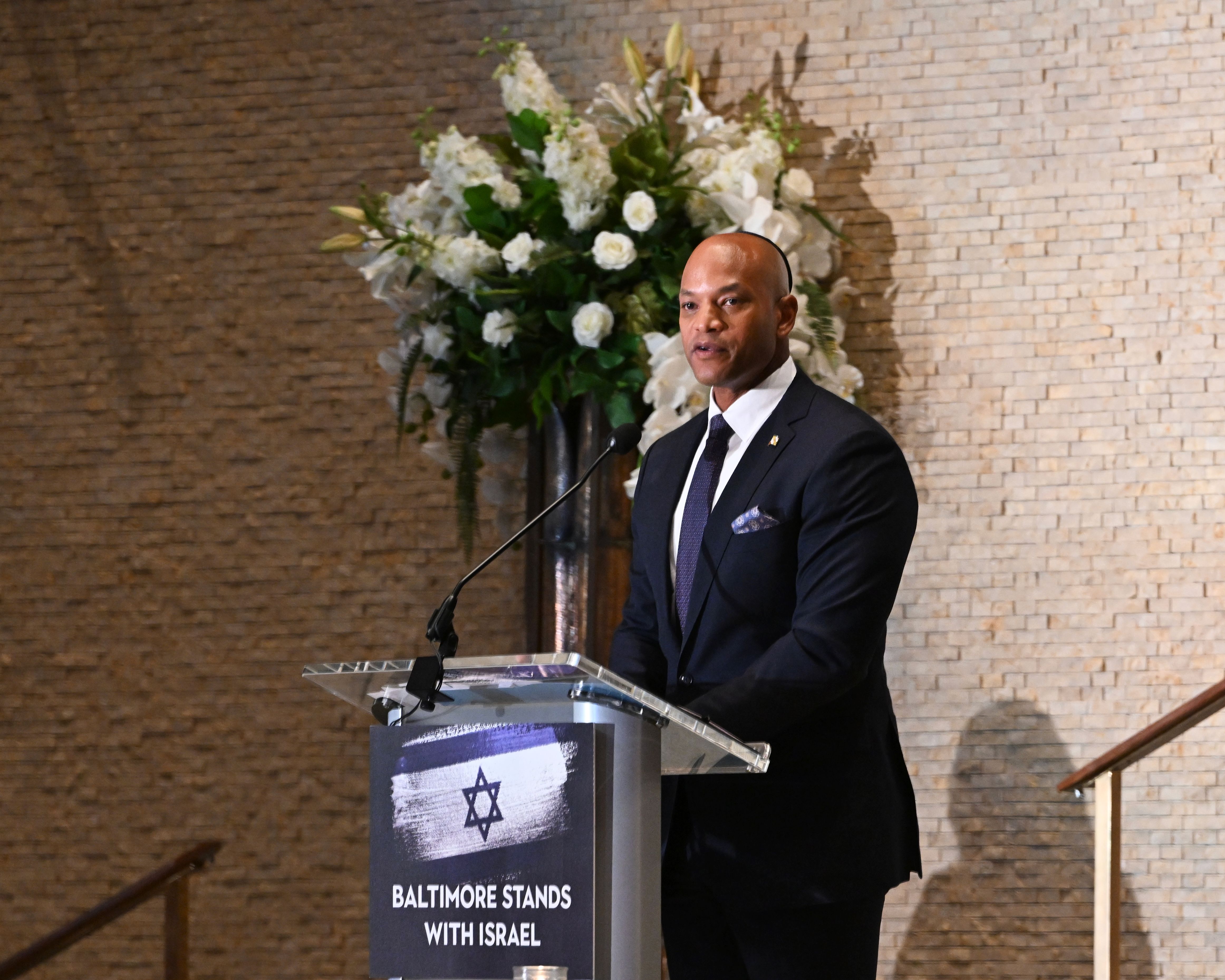 A man in a dark suit speaks at a podium bearing a Star of David and solidarity sign.