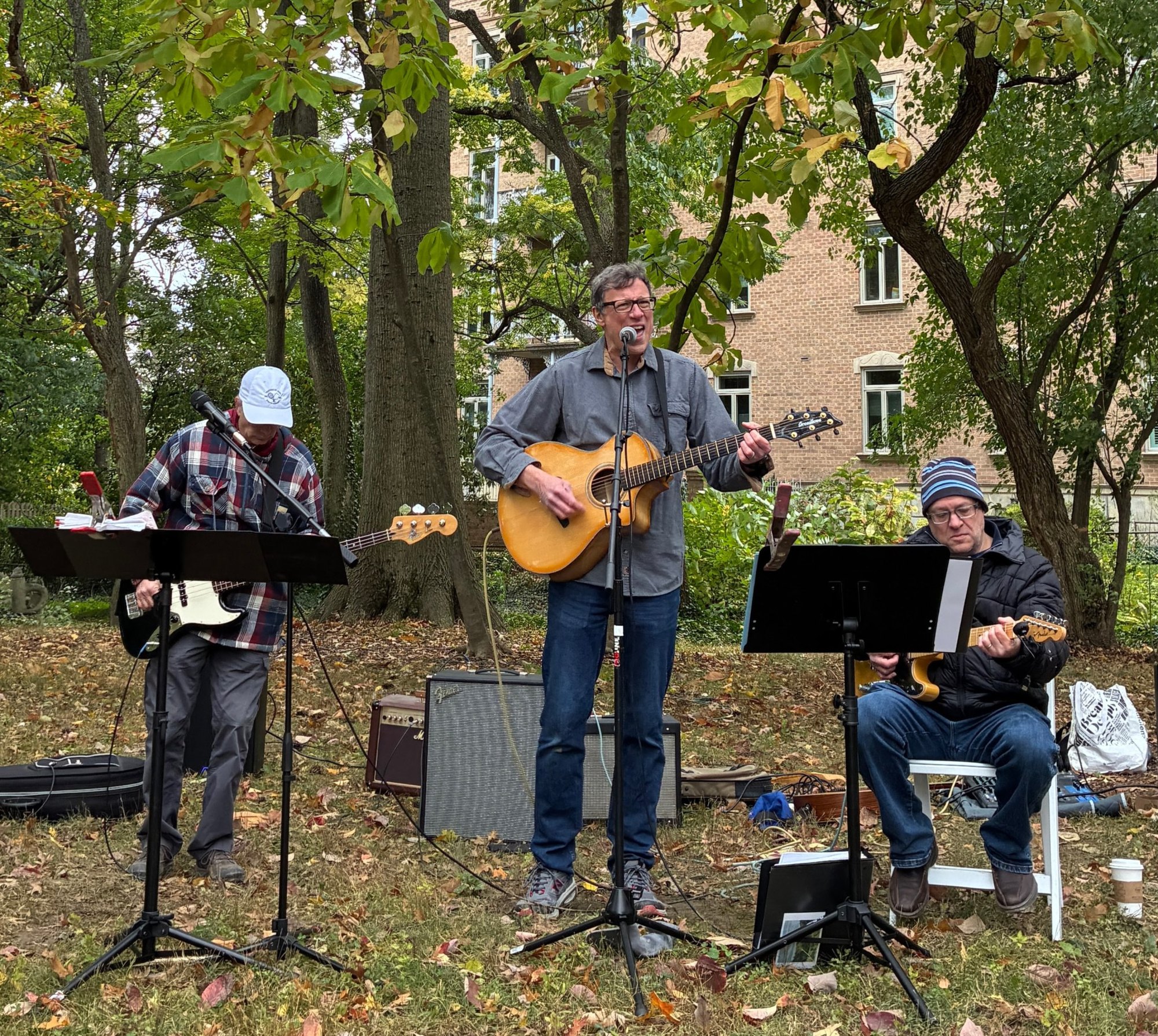 Three musicians perform outdoors on a leaf-covered lawn surrounded by autumn trees.